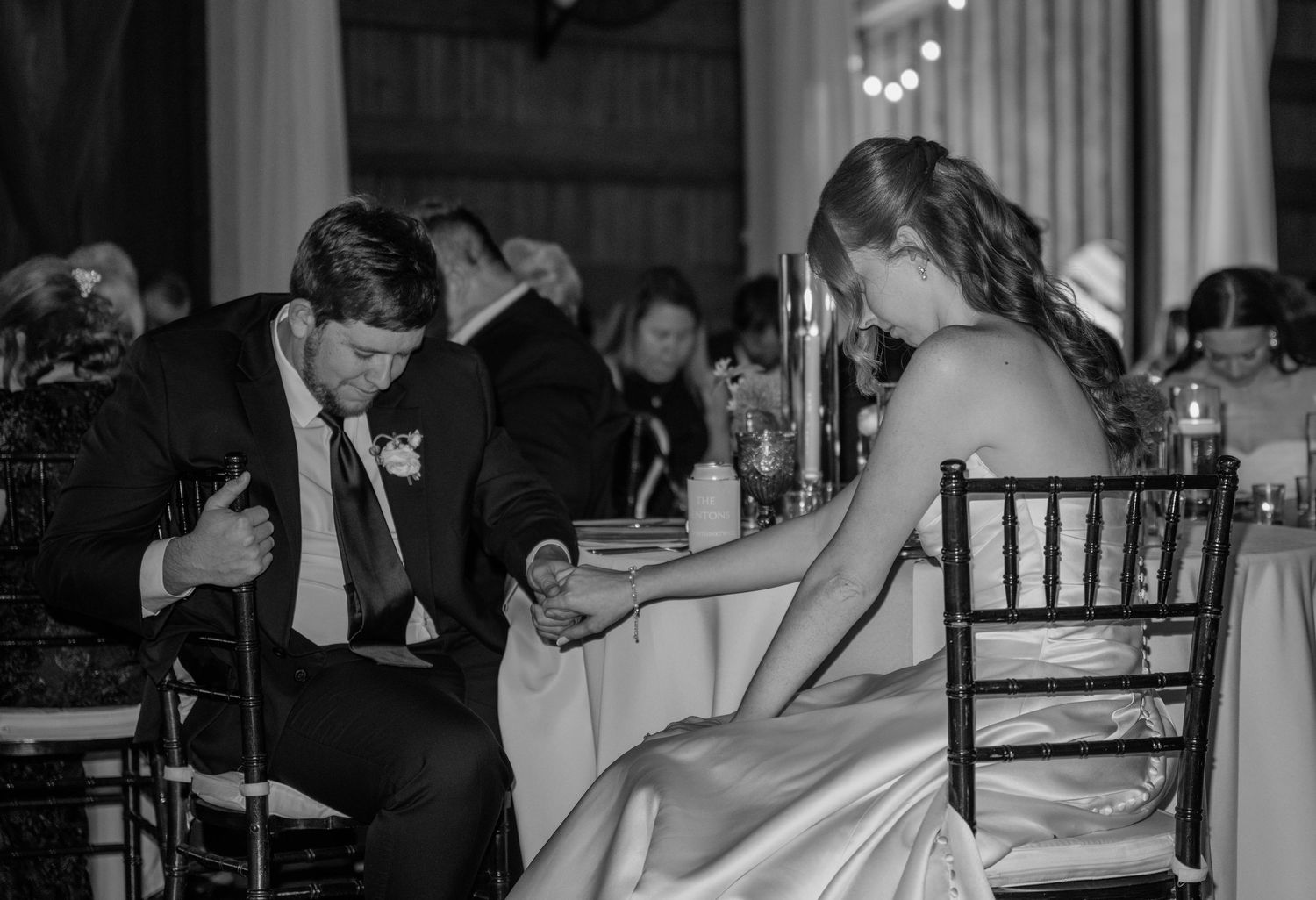 Bride and groom hold hands at a reception table while the groom bows his head during a quiet moment in a black and white photo.