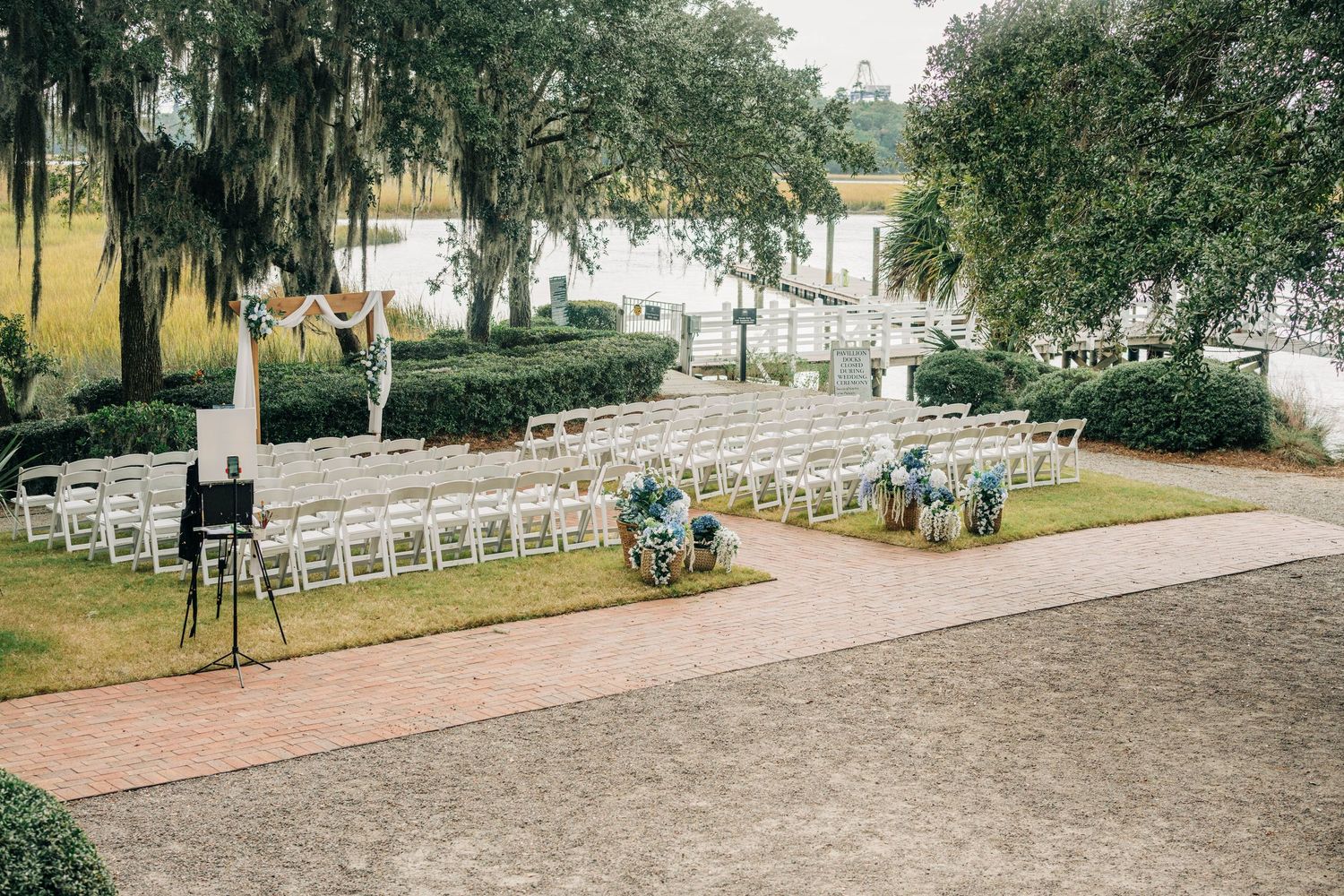 White chairs line an outdoor wedding ceremony site under mossy oak trees with a waterfront view and a brick pathway.