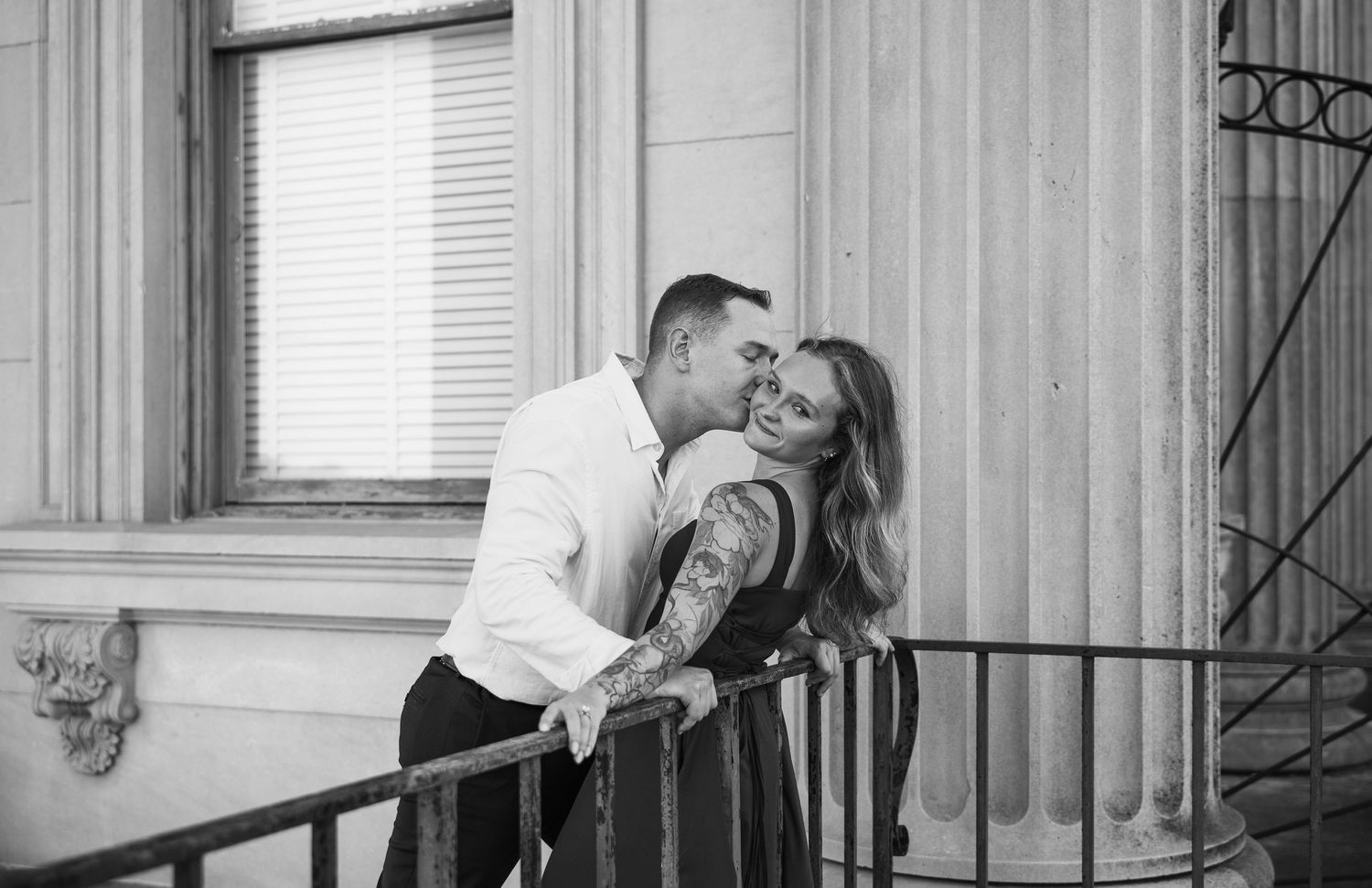 Black and white engagement photo of a tattooed woman being kissed on the cheek by her fiancé on a historic building's iron staircase.