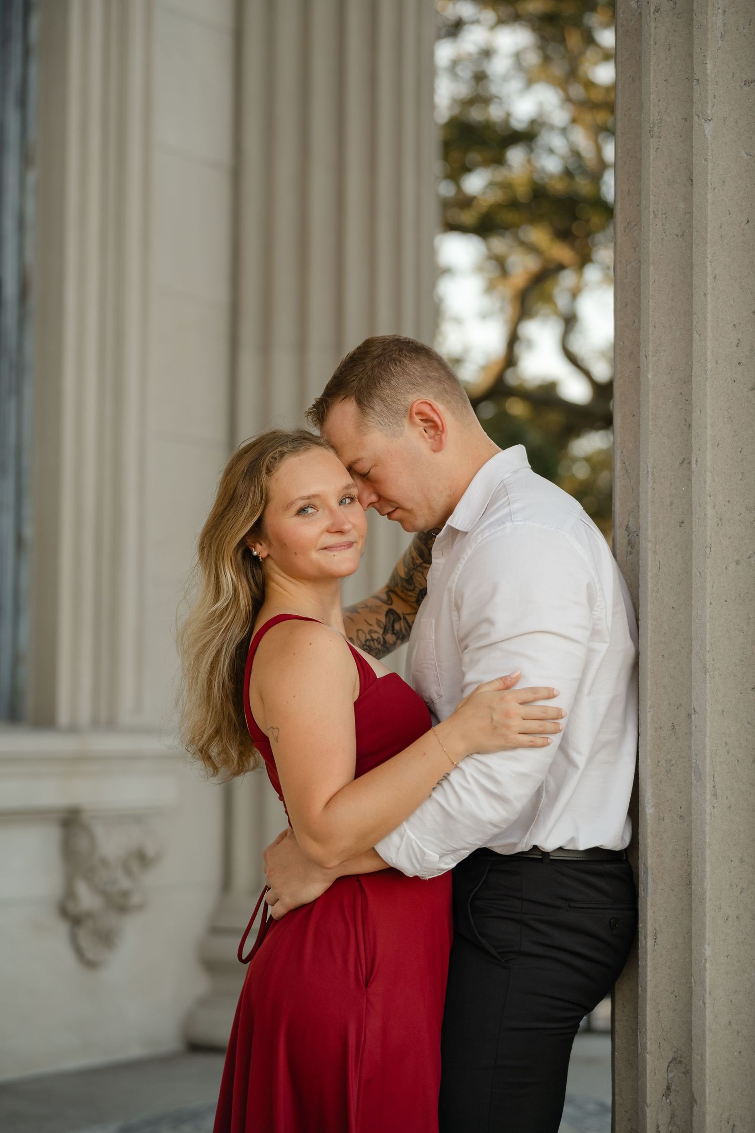Woman in a red dress smiles at the camera as her partner in a white shirt rests his forehead on her shoulder beside stone columns.