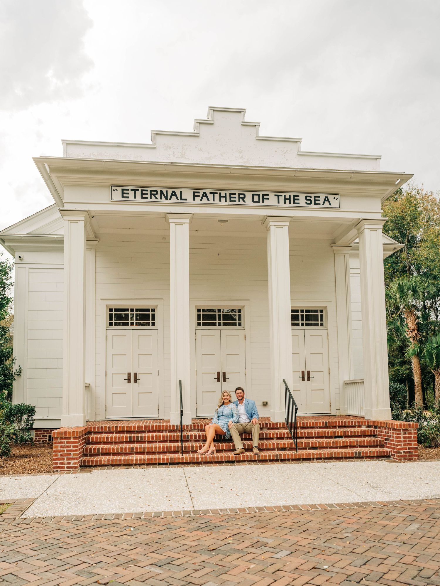 Engaged couple sitting on brick steps in front of a white chapel reading Eternal Father of the Sea.