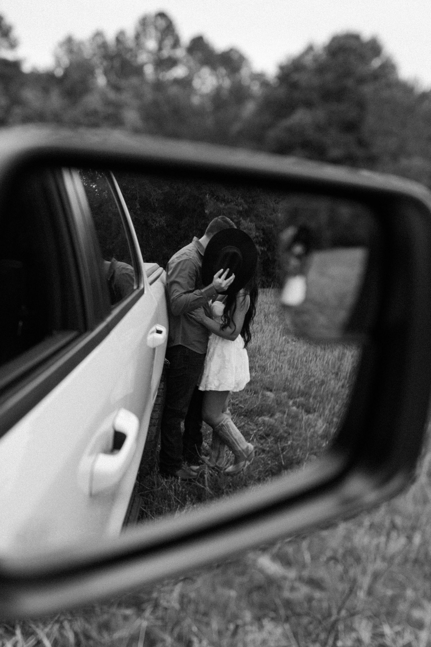 Black and white side mirror reflection of a couple embracing beside a truck in a grassy field.