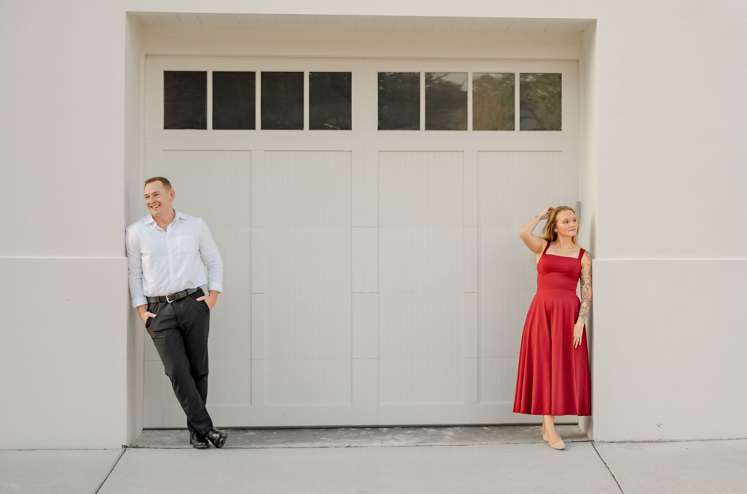 Engagement portrait of a man in a white shirt and a woman in a red dress standing apart beside a cream garage door.