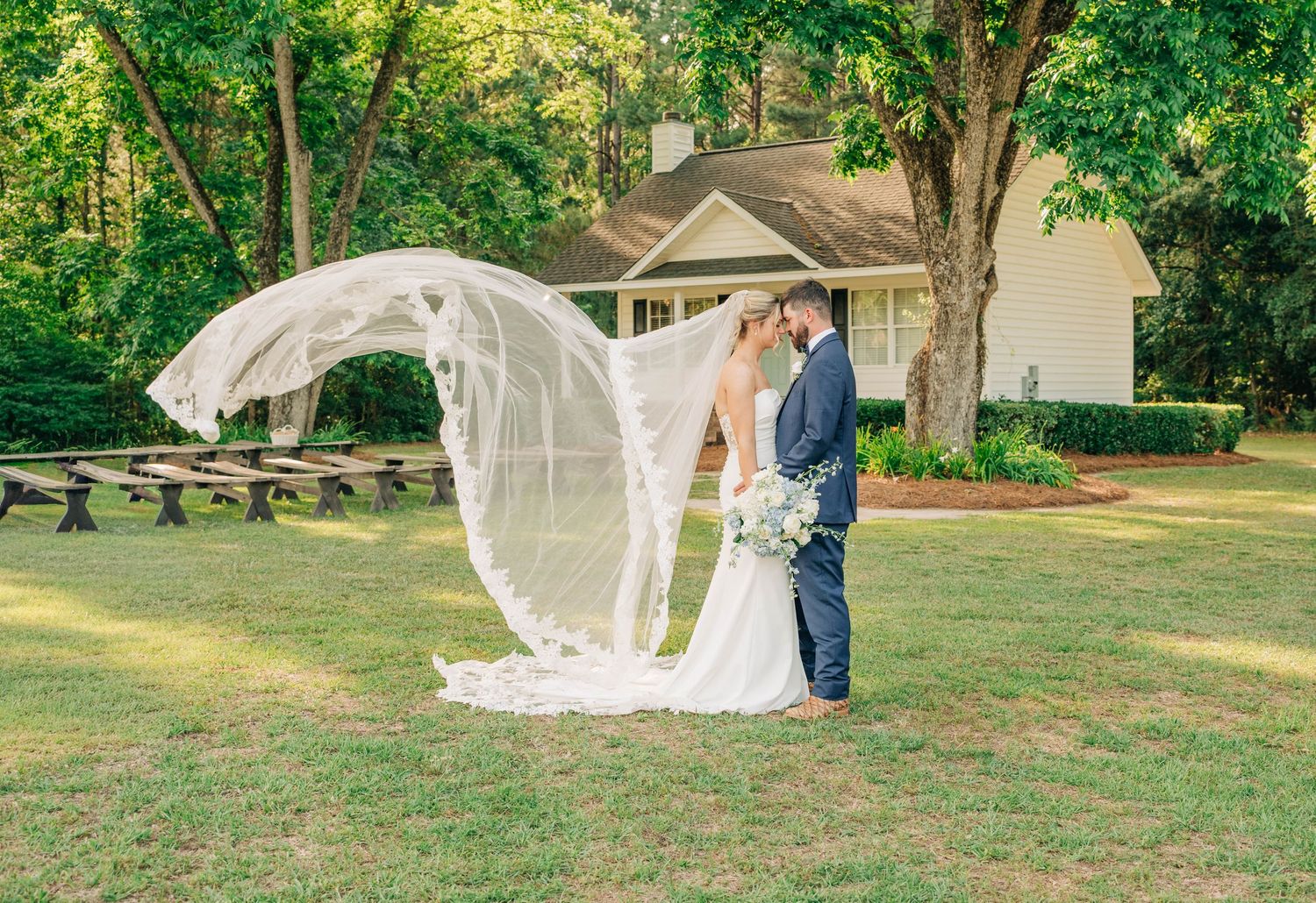 A bride and groom embrace under a tree as her long sheer veil billows in the wind, with a white cottage and wooden benches behind them.