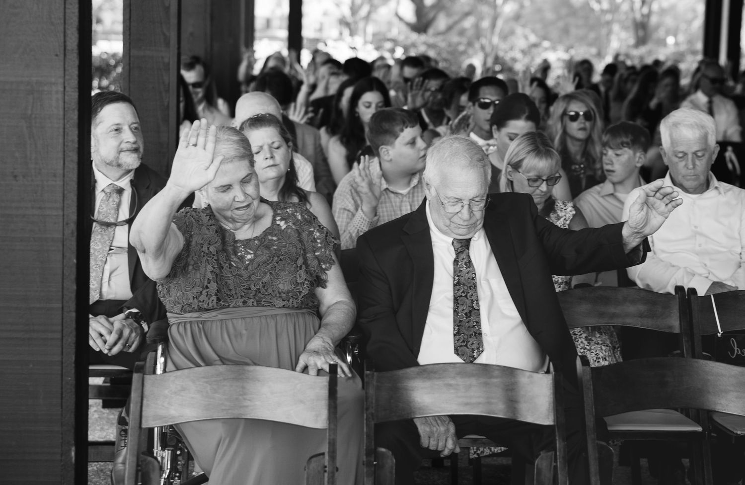 Black and white photo of an elderly woman raising her hand in prayer beside a man in a suit among wedding guests seated outdoors.