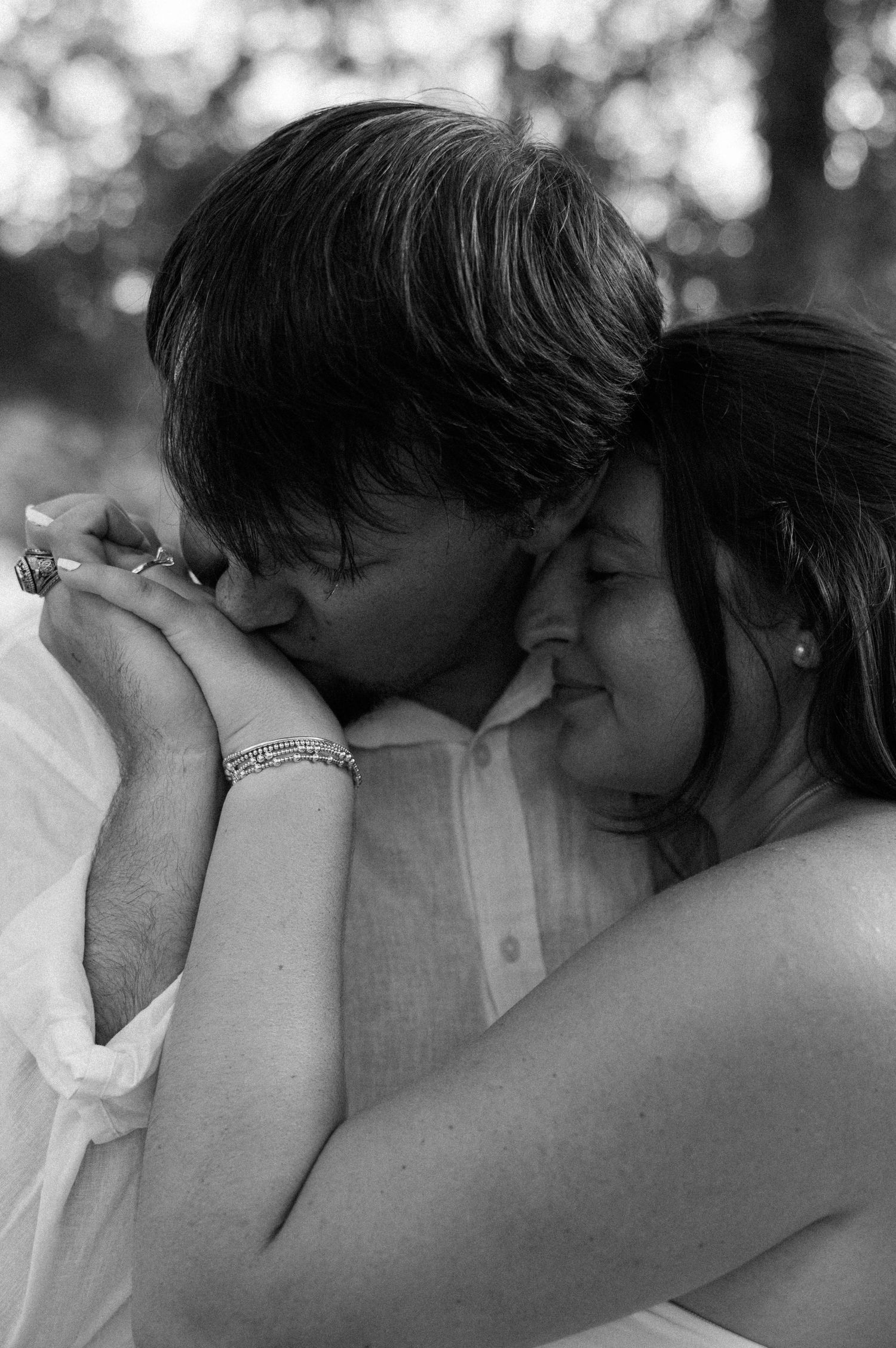 Black and white portrait of a couple embracing as the man kisses the woman's hand, showing her ring and bracelet.