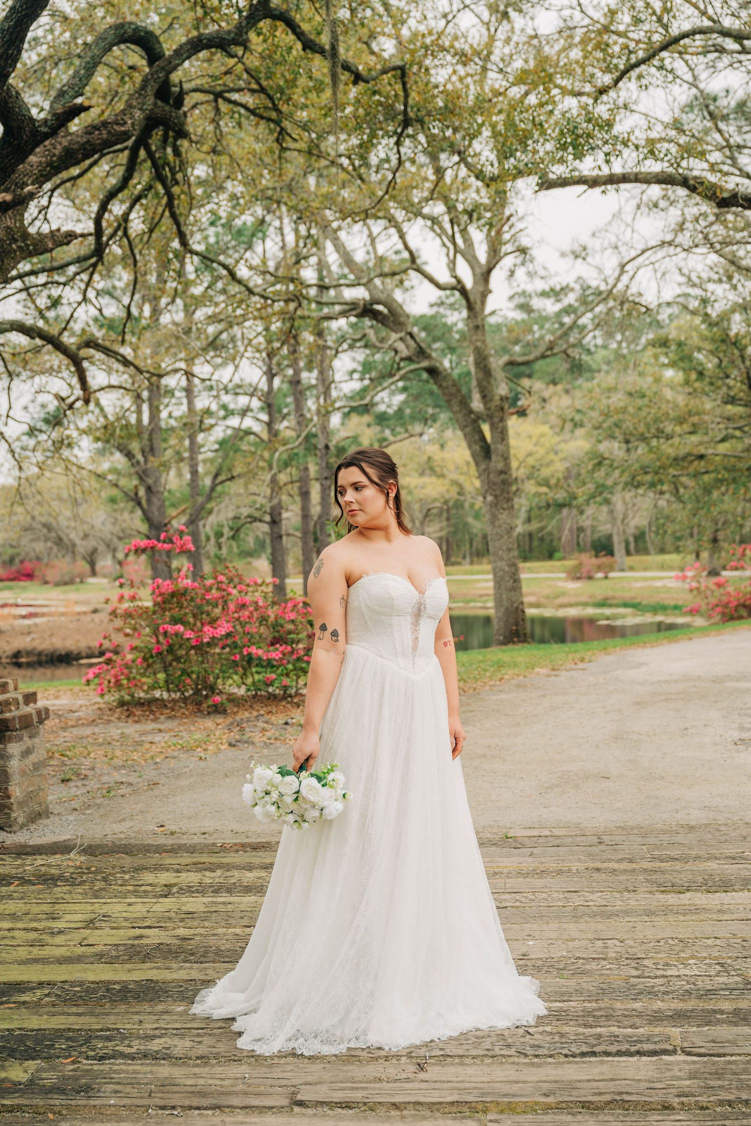 Bride in a beaded A-line tulle gown holds a white bouquet on a wooden boardwalk near blooming pink azaleas.