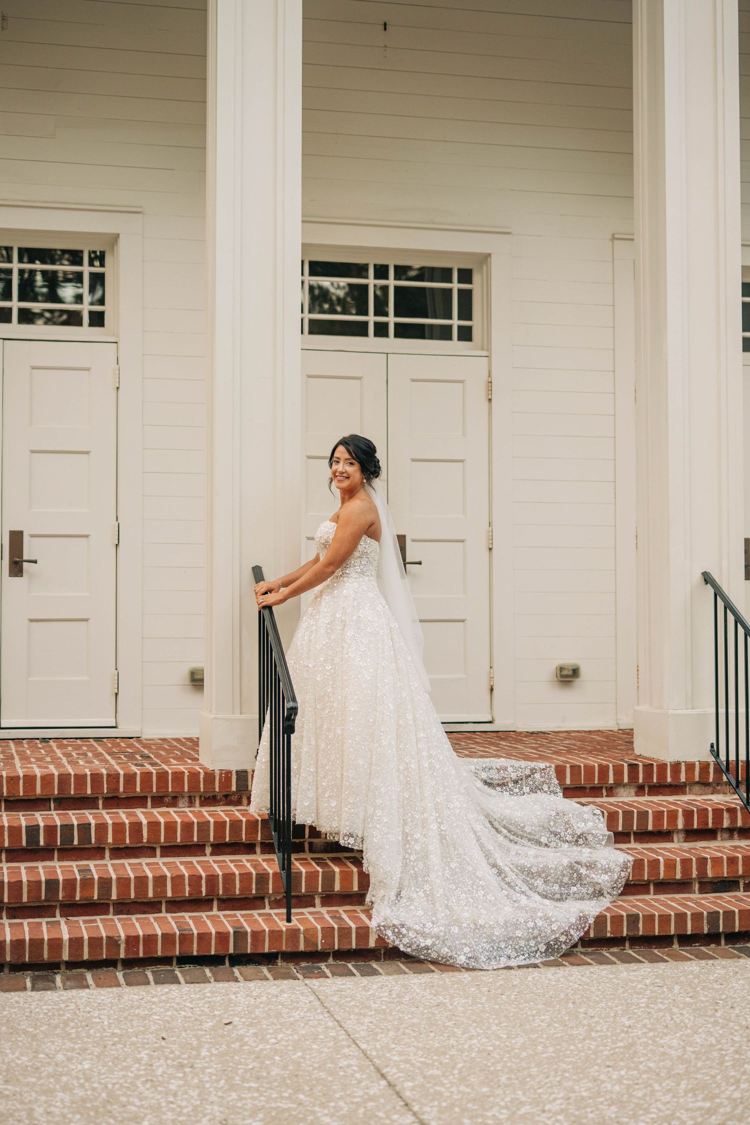 Smiling bride in a sparkling tulle gown with a long train stands on red brick steps beside a white columned building.