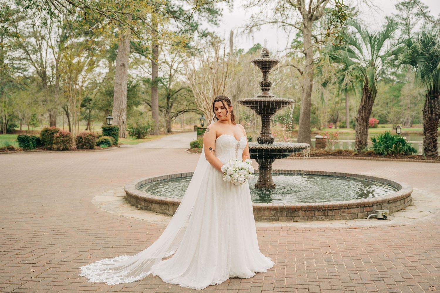 Bride in a flowing tulle wedding dress holds white flowers beside a tiered stone fountain in a brick garden courtyard.