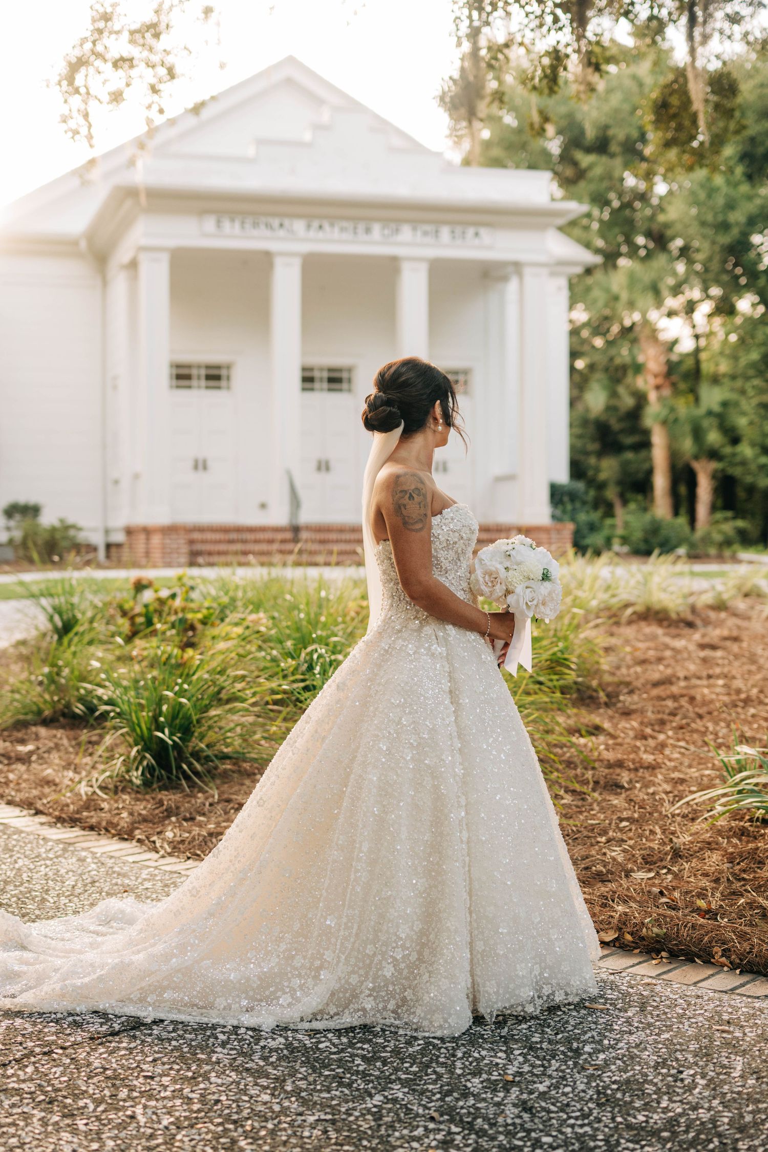 Bride with tattooed arm and updo holds a white bouquet in a glittering gown outside a white chapel with columns.