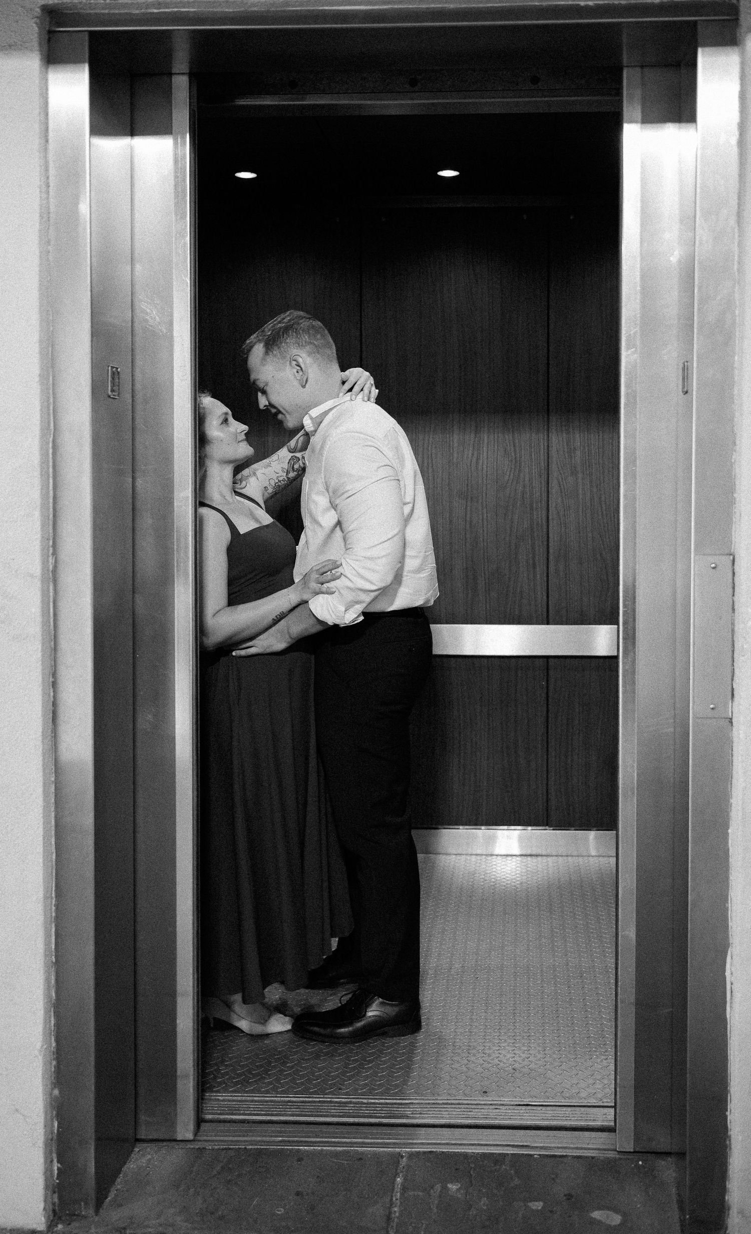 Black and white engagement photo of a couple embracing inside an elevator with polished metal doors.