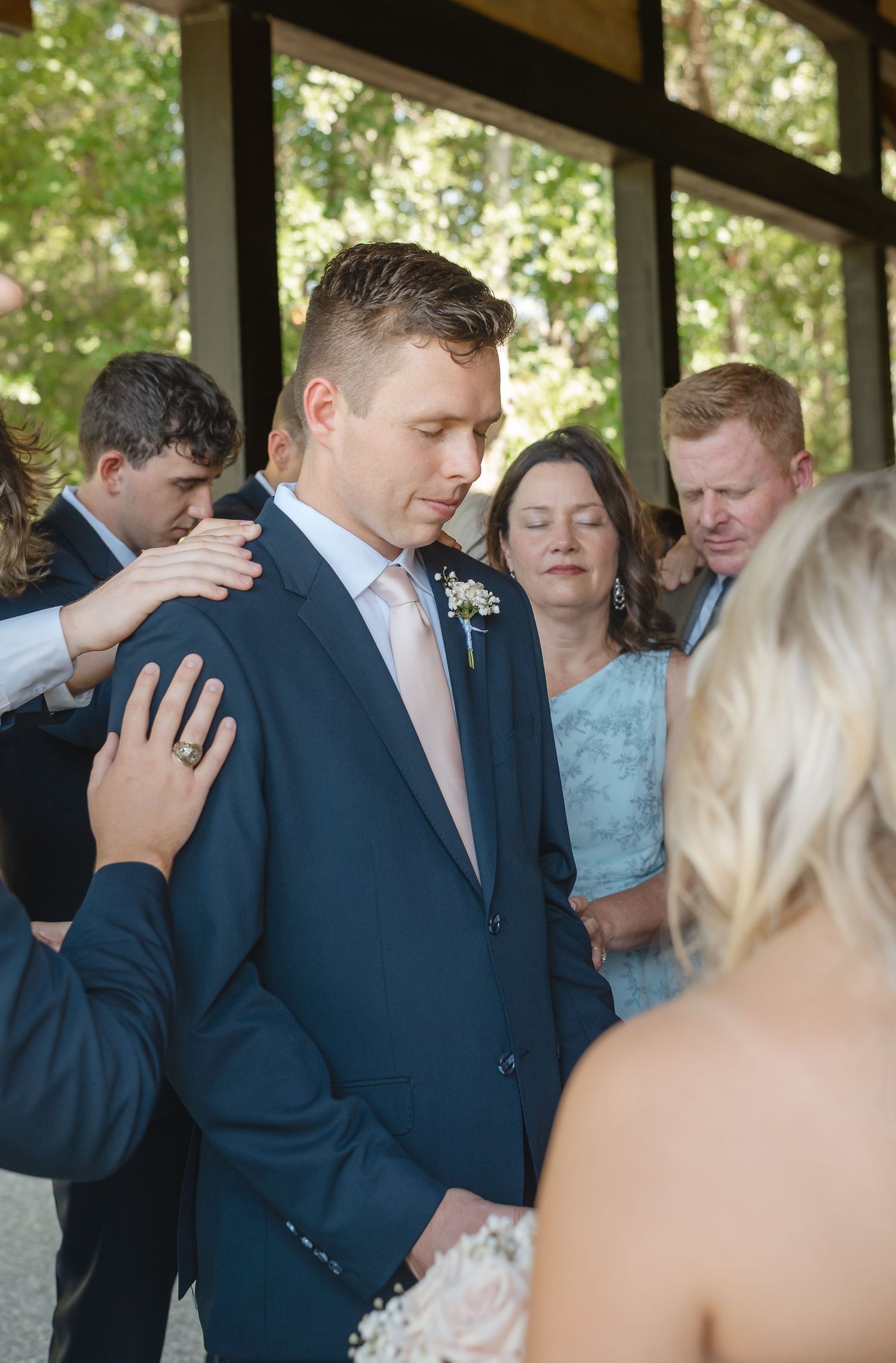 Groom in a navy suit bows his head during a wedding prayer as loved ones touch his shoulders and guests stand reverently nearby.