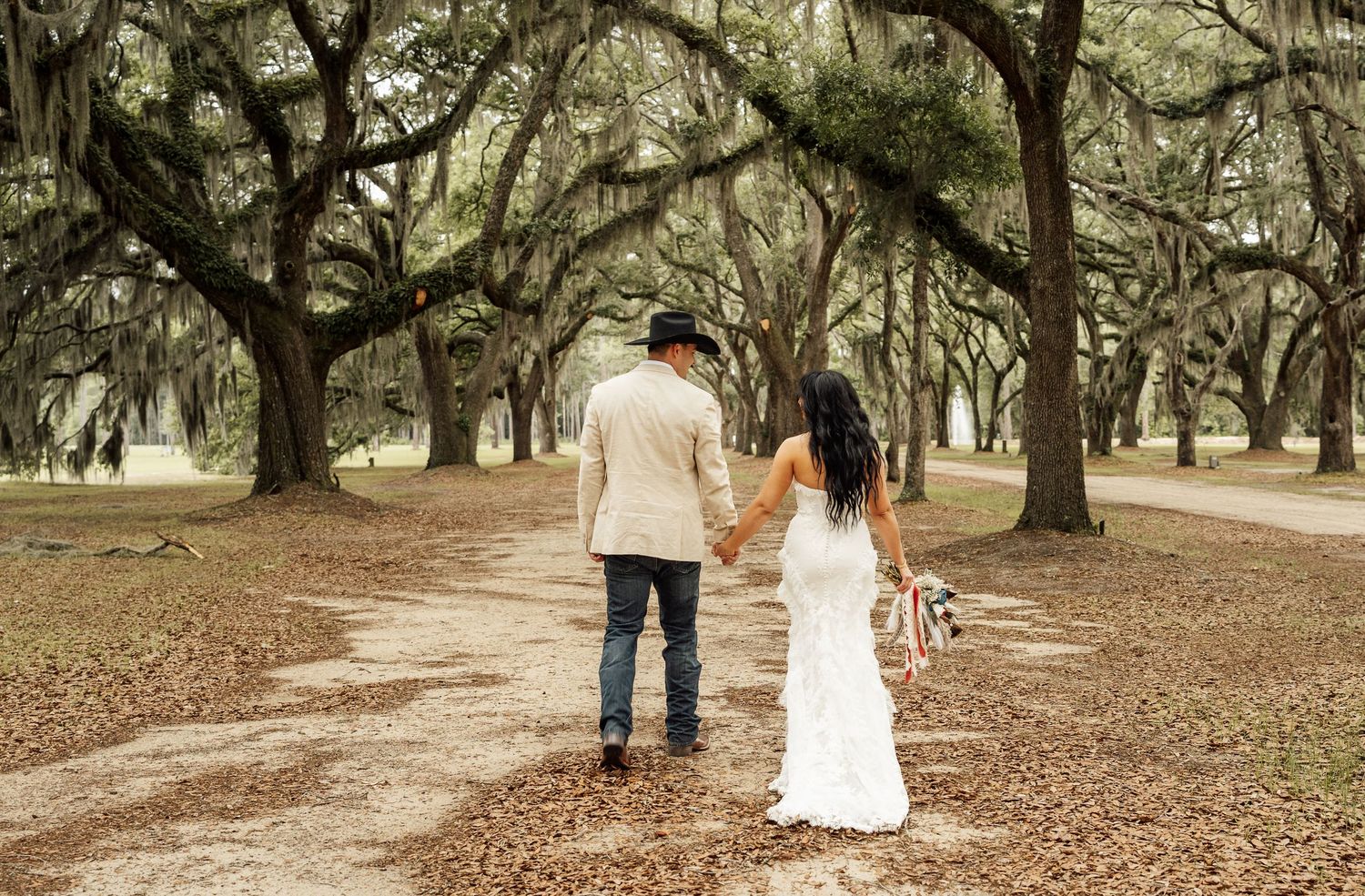 Bride and groom in a cowboy hat walk hand in hand down a dirt path lined with moss-covered oak trees on their wedding day.