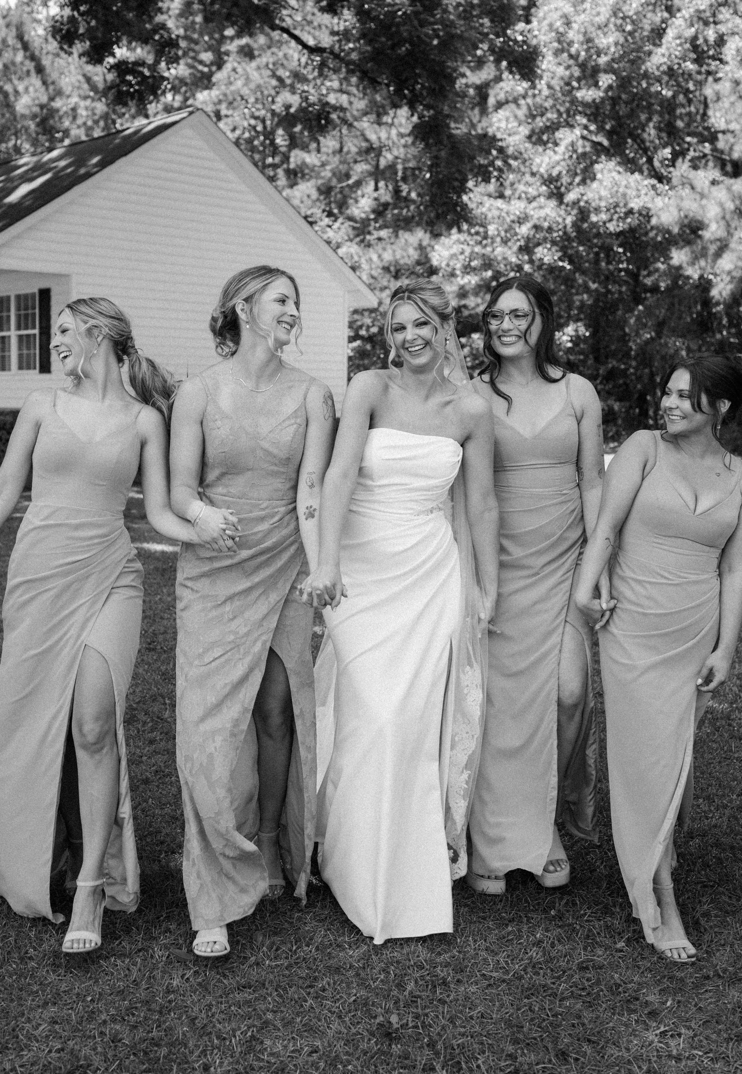 A bride in a white strapless gown walks arm in arm with four smiling bridesmaids in slit dresses in a black and white outdoor photo.