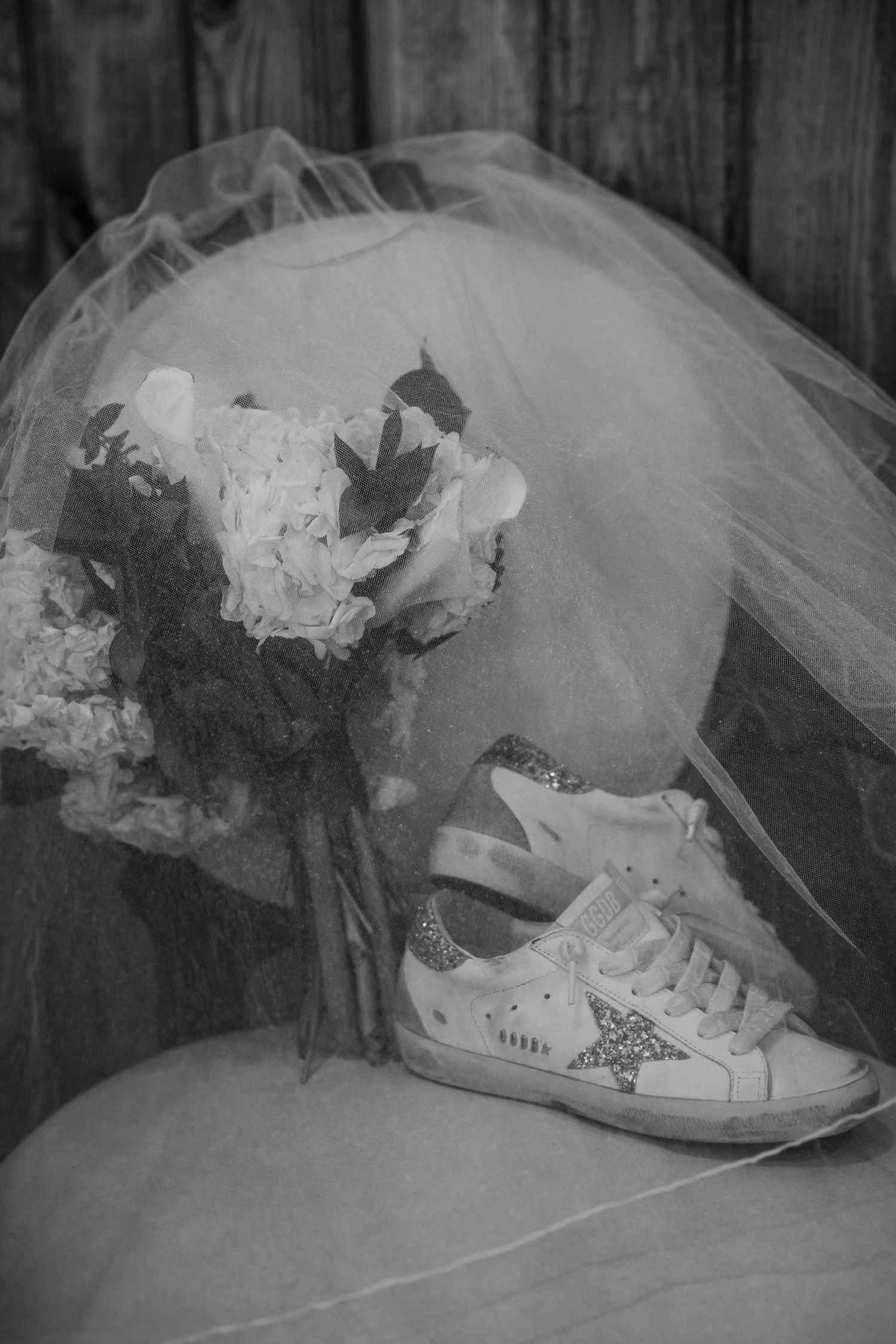 Black and white detail shot of a bridal bouquet, flowing veil and glitter star wedding sneakers resting on a chair.
