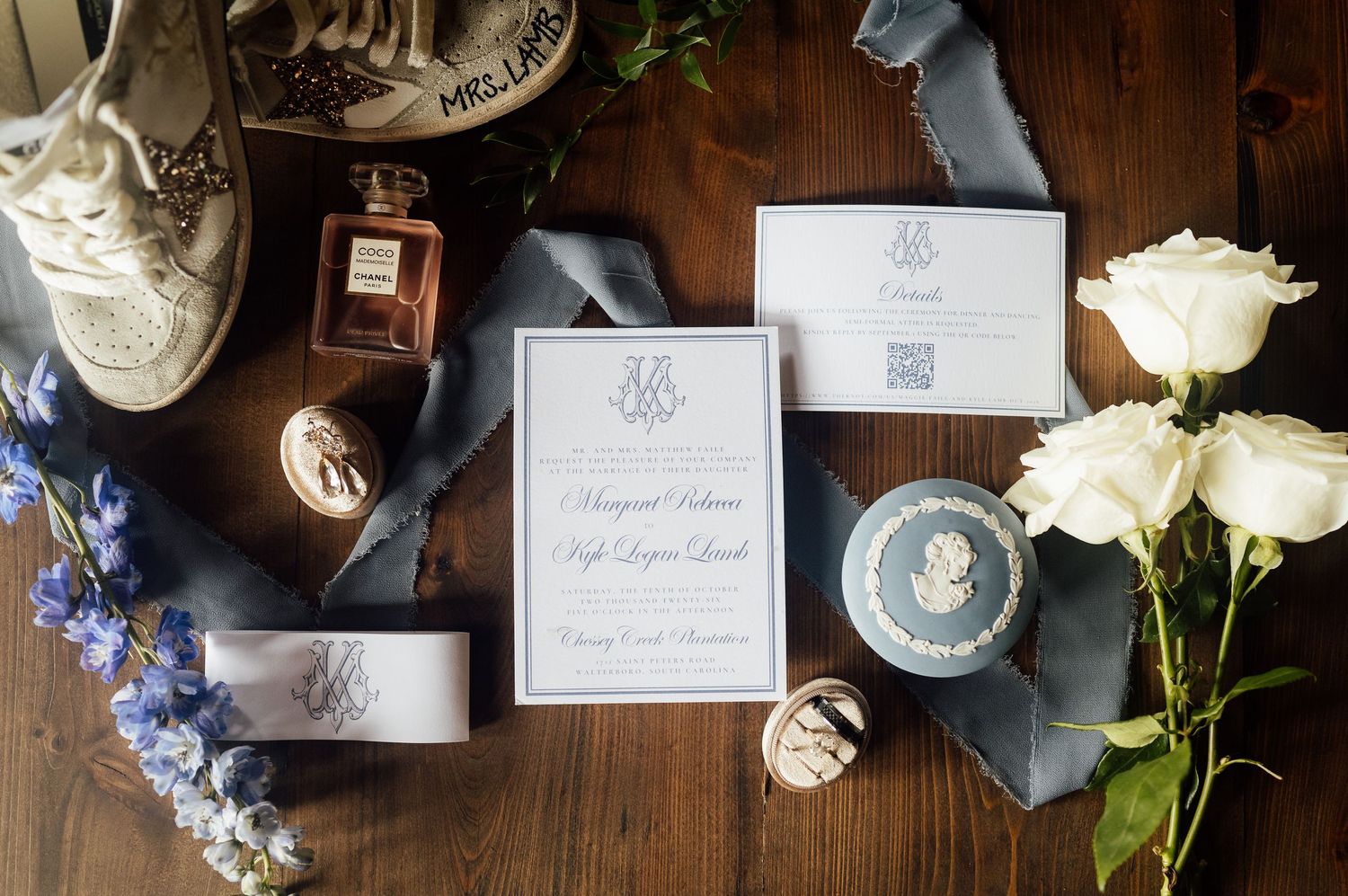 Flat lay of a wedding invitation suite with blue ribbon, perfume bottle, white roses, rings and sneakers on a wooden table.