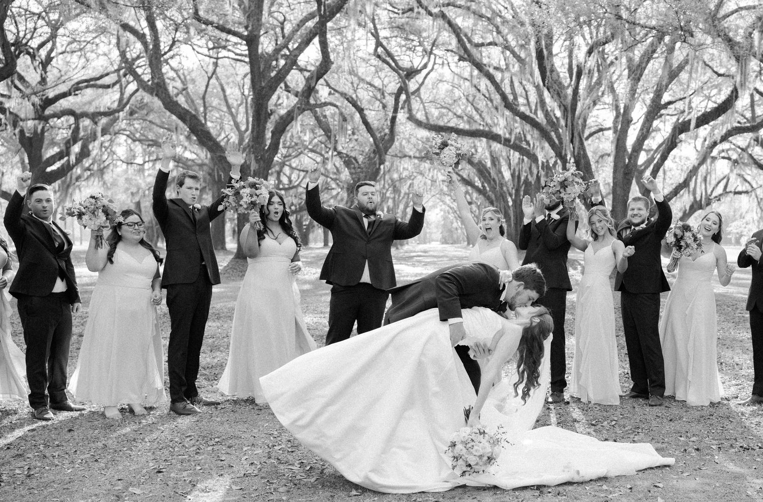 Bride and groom share a dipped kiss under oak trees as wedding guests in formal attire cheer with raised arms behind them.