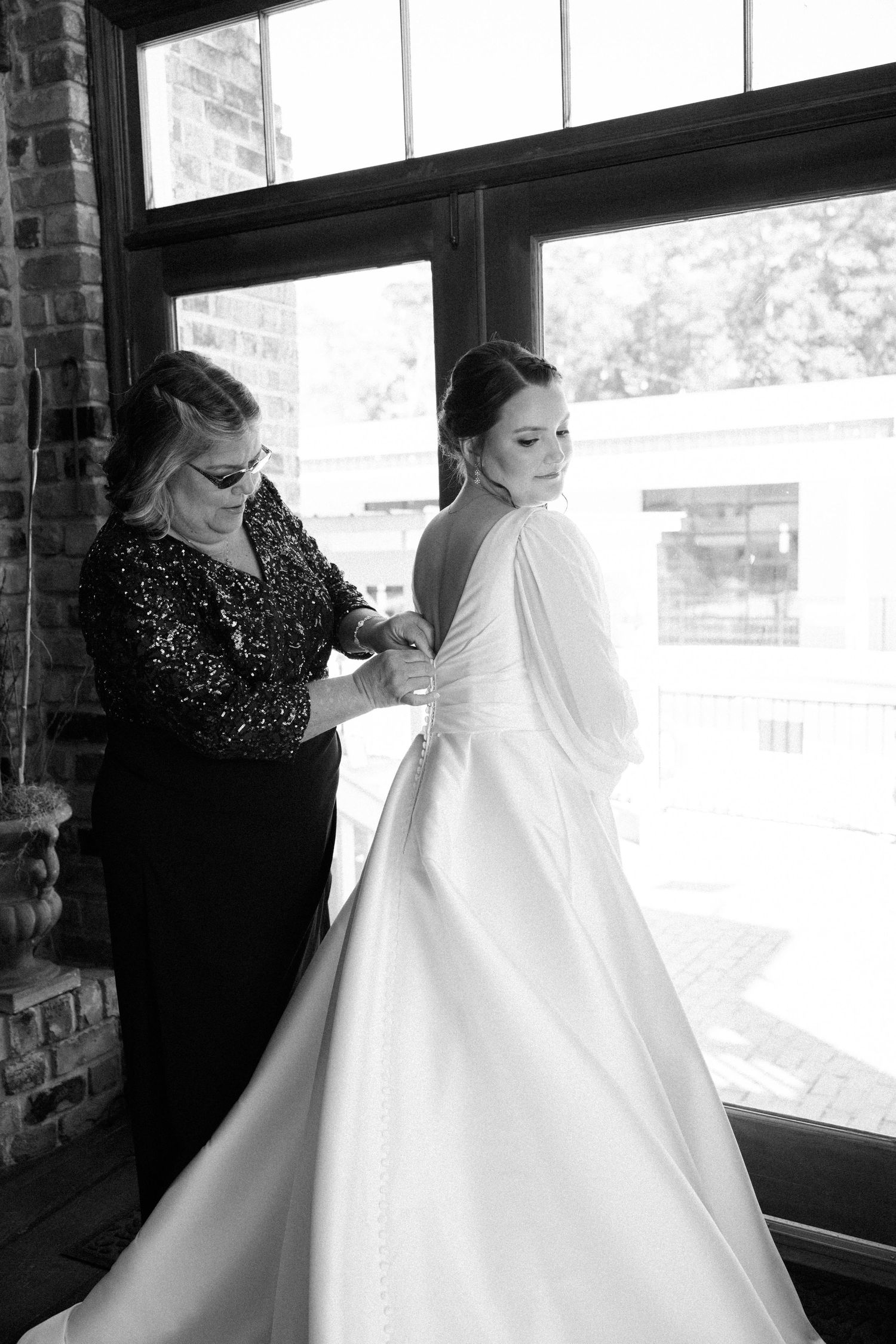 Black and white photo of a mother in a sequined top buttoning the back of her daughter's wedding gown by a large window.
