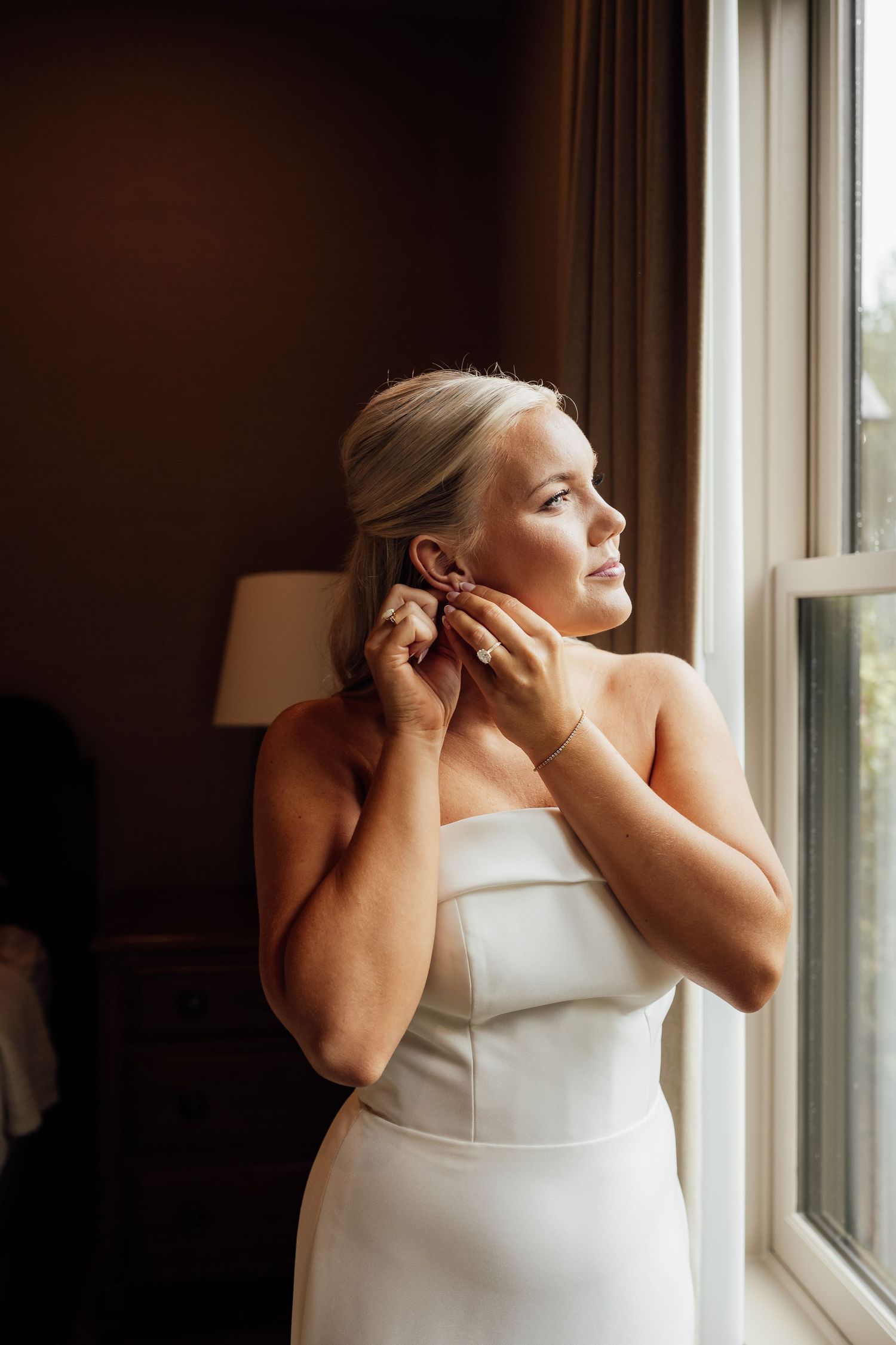 Bride in a strapless white gown looks out a window while putting on an earring in soft natural light during wedding preparations.