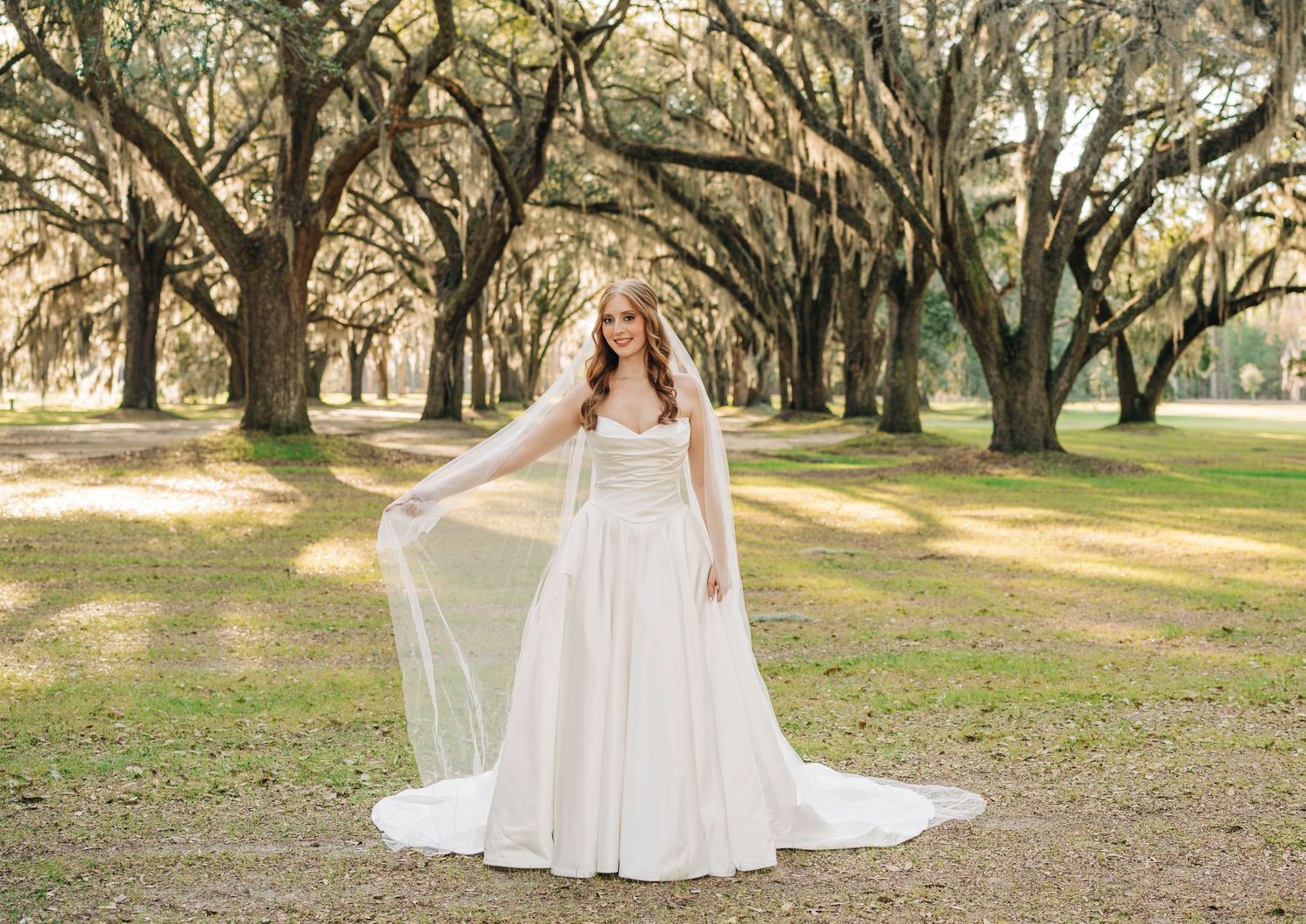 Bride in an elegant ivory gown lifting her sheer veil while standing on a grassy lawn under mossy oak trees.