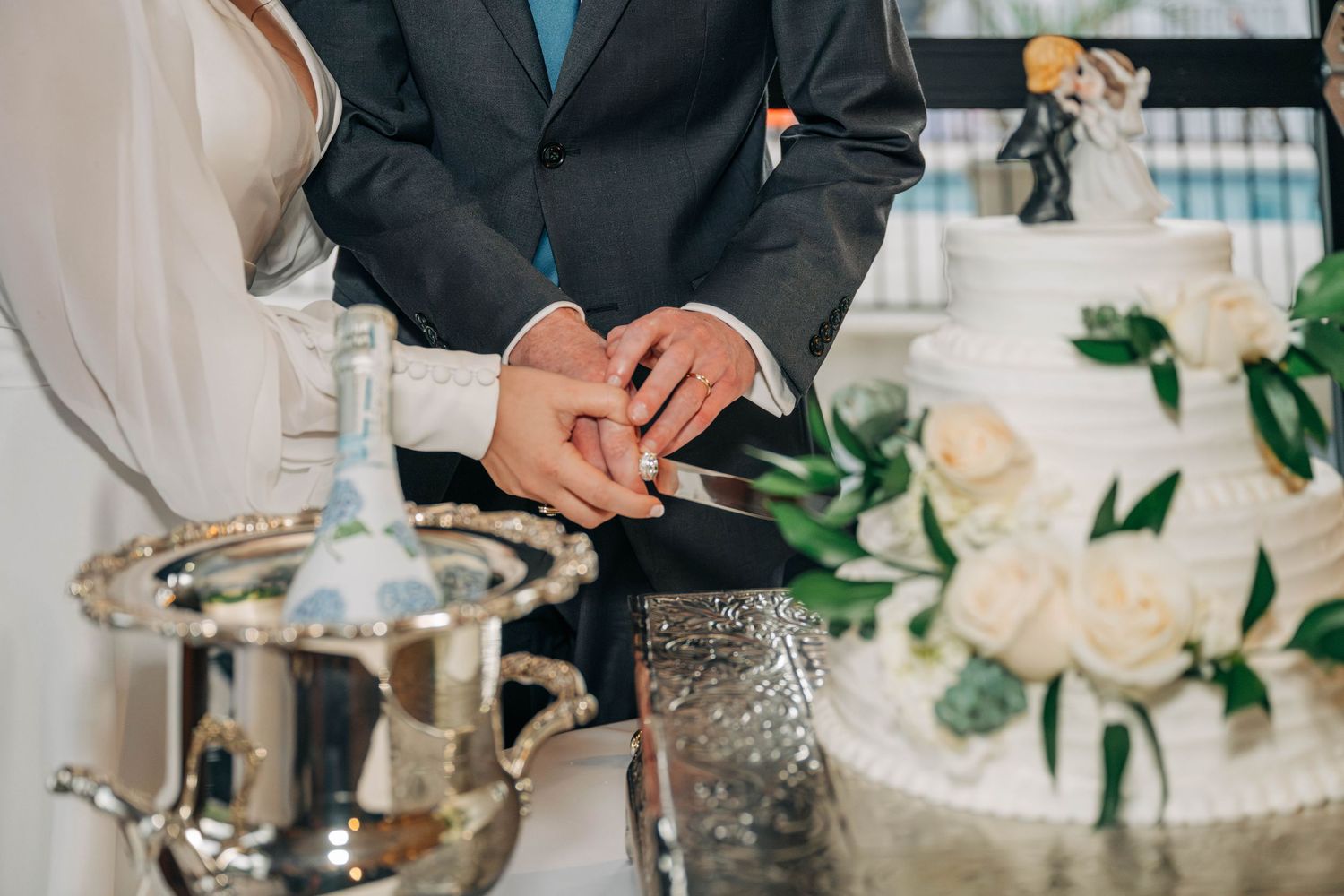 A bride and groom hold hands as they cut their white wedding cake decorated with cream roses and greenery.