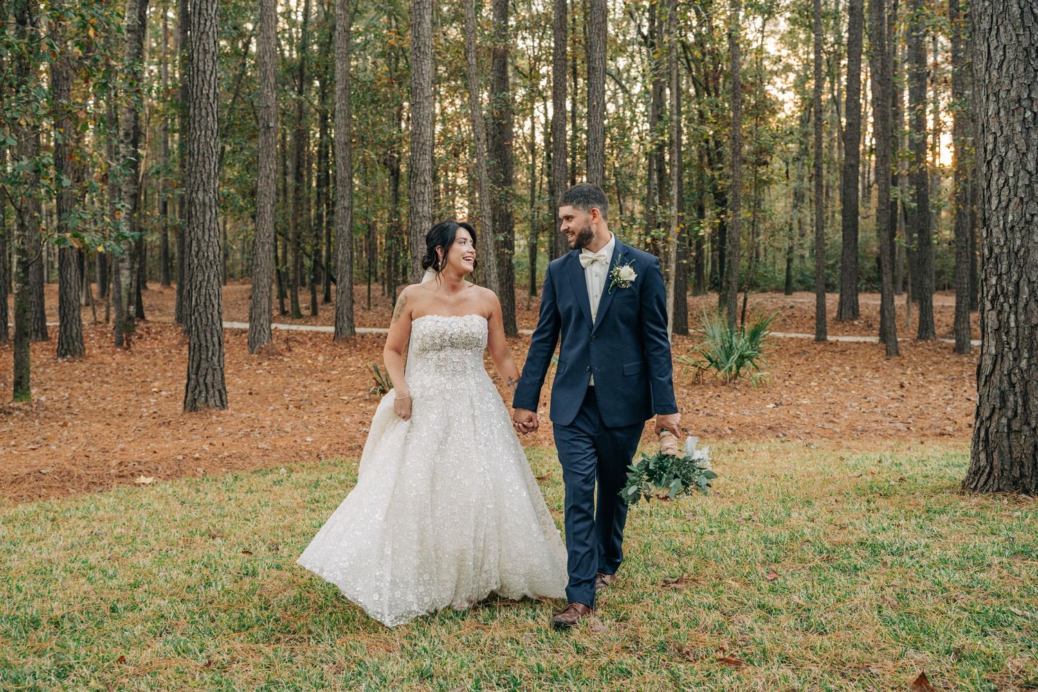 Smiling bride in a strapless gown and groom in a navy suit hold hands and walk through a sunlit pine forest with a bouquet.