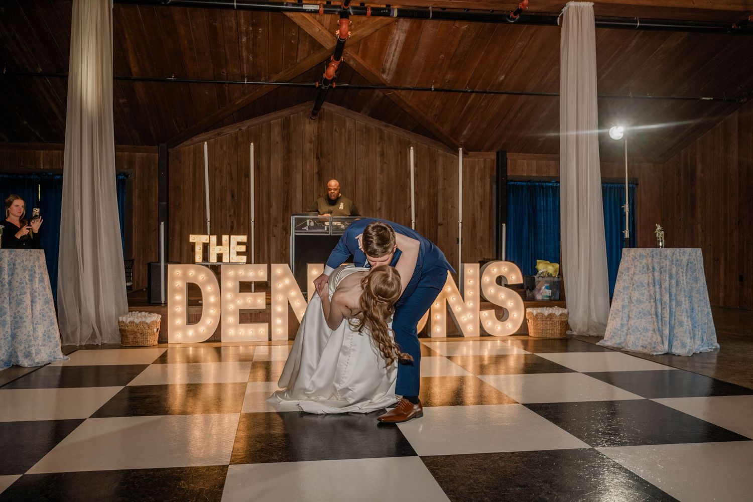 Newlyweds share a dramatic dance dip on a checkered dance floor in front of large illuminated marquee letters at their reception.