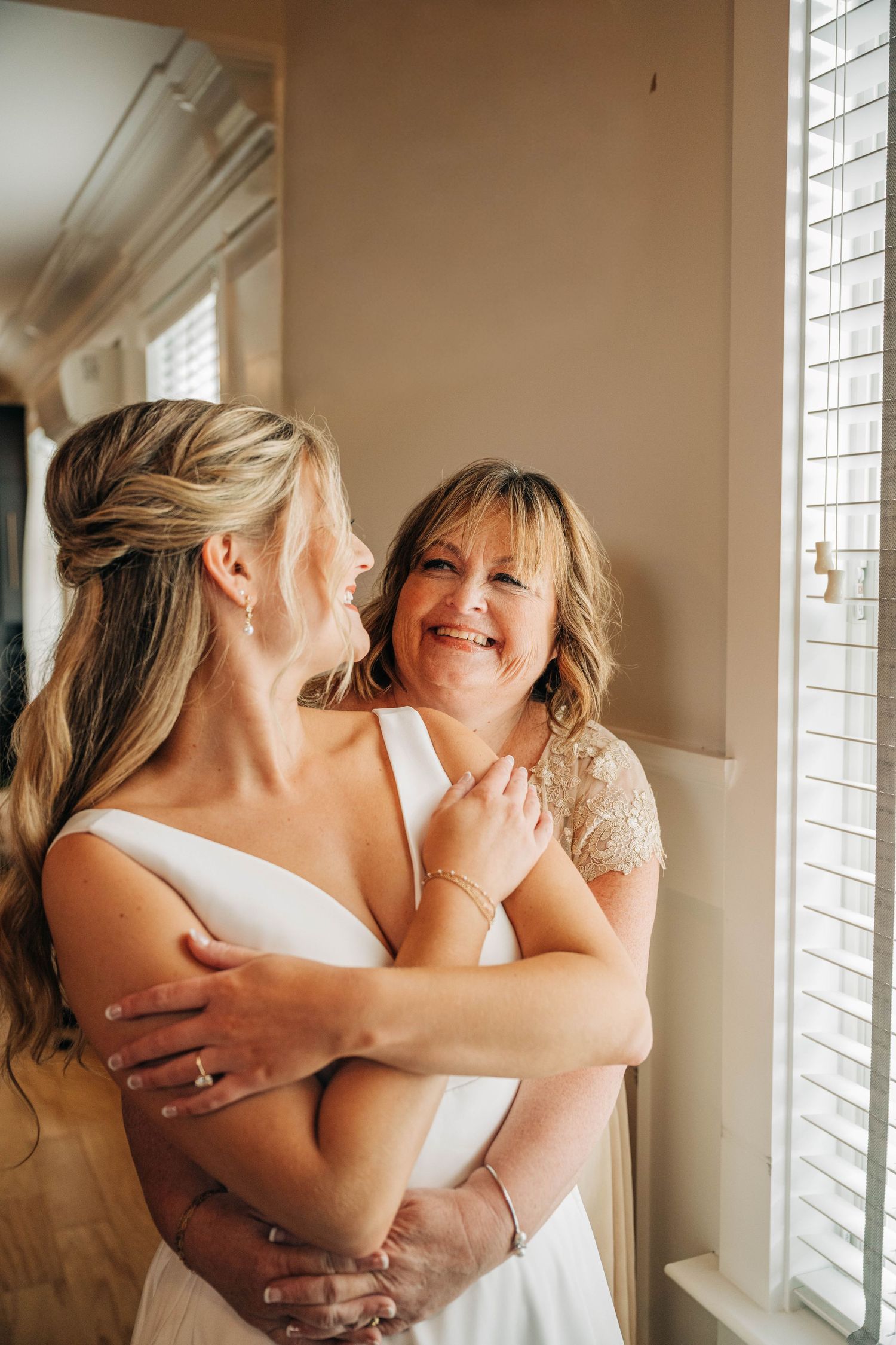 A bride in a white dress shares a joyful hug with her smiling mother beside a sunlit window on the wedding day.