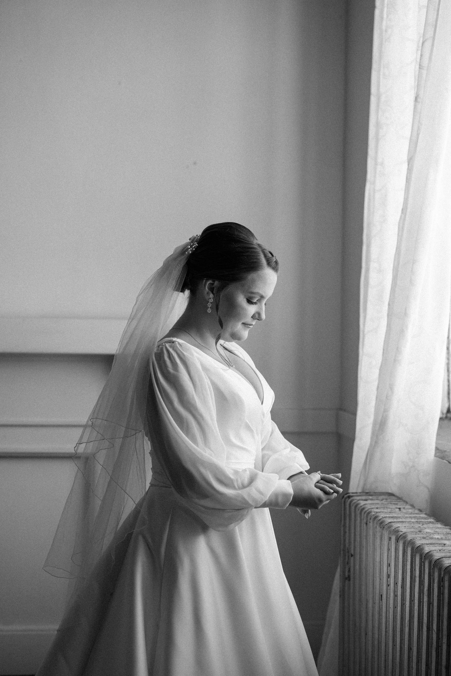 Black and white portrait of a bride in a long-sleeved gown and veil looking down at her hands beside a sunlit window.