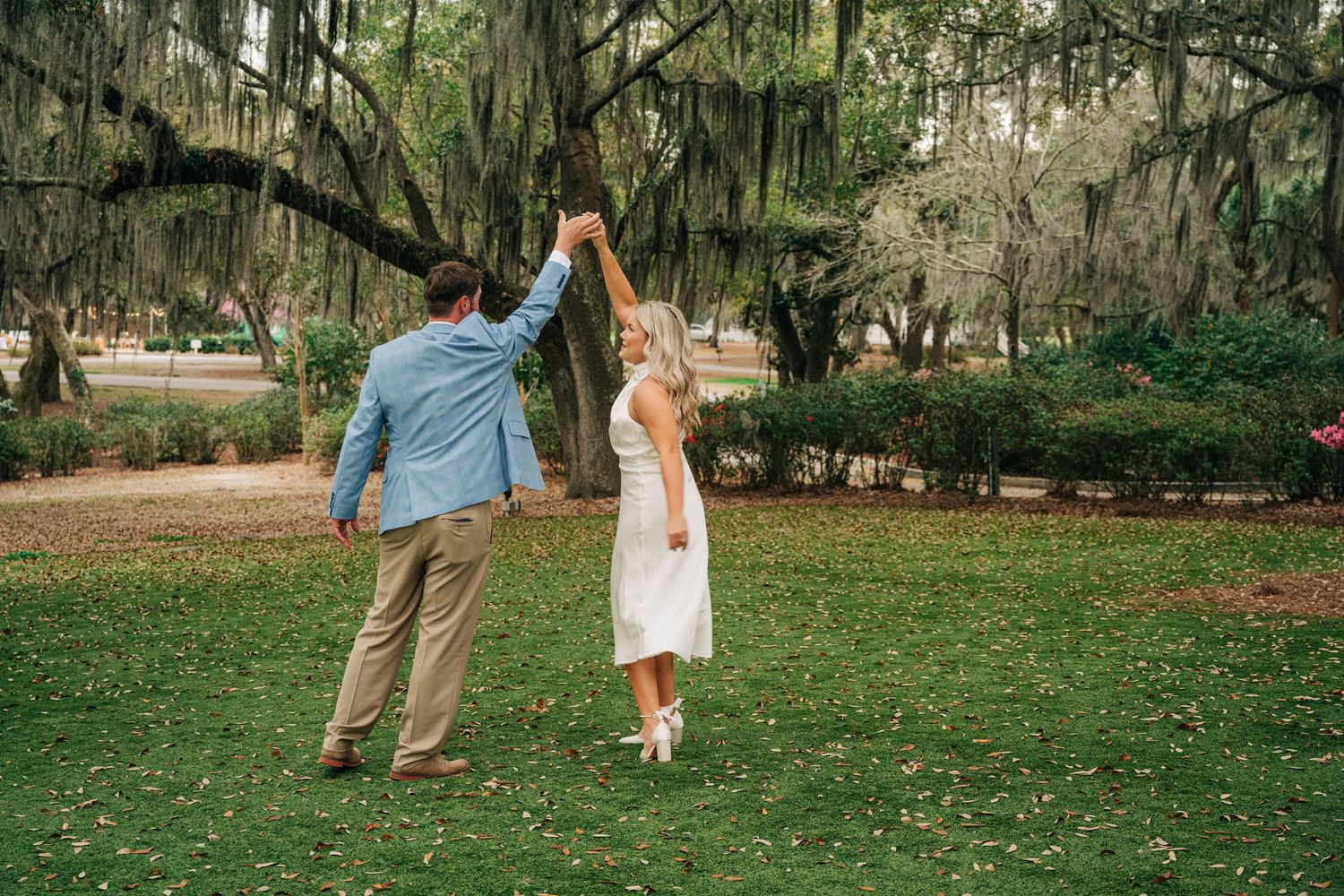Couple dancing a twirl on a lawn beneath oak trees draped in Spanish moss during an engagement photo session.