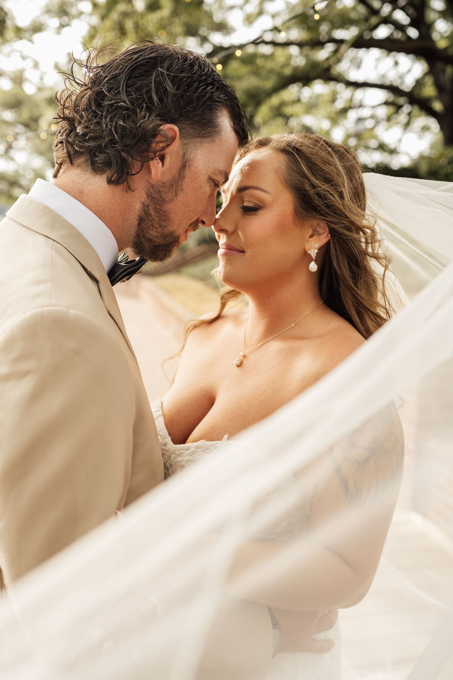 Bride and groom touch foreheads in a tender moment as a sheer wedding veil drifts around them outdoors.