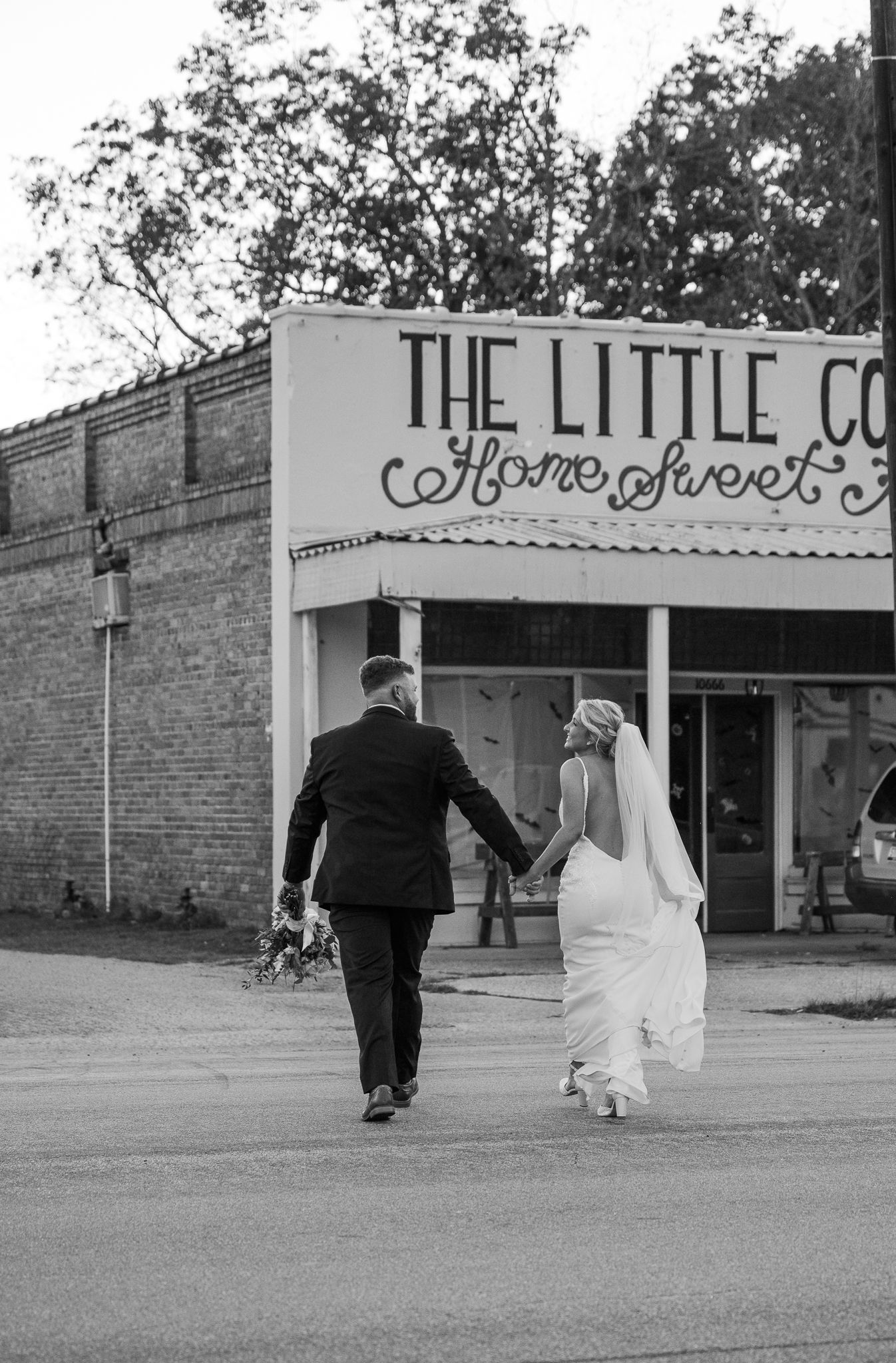 Black and white photo of a bride in a veil and groom holding hands as they walk toward a vintage storefront reading Home Sweet.
