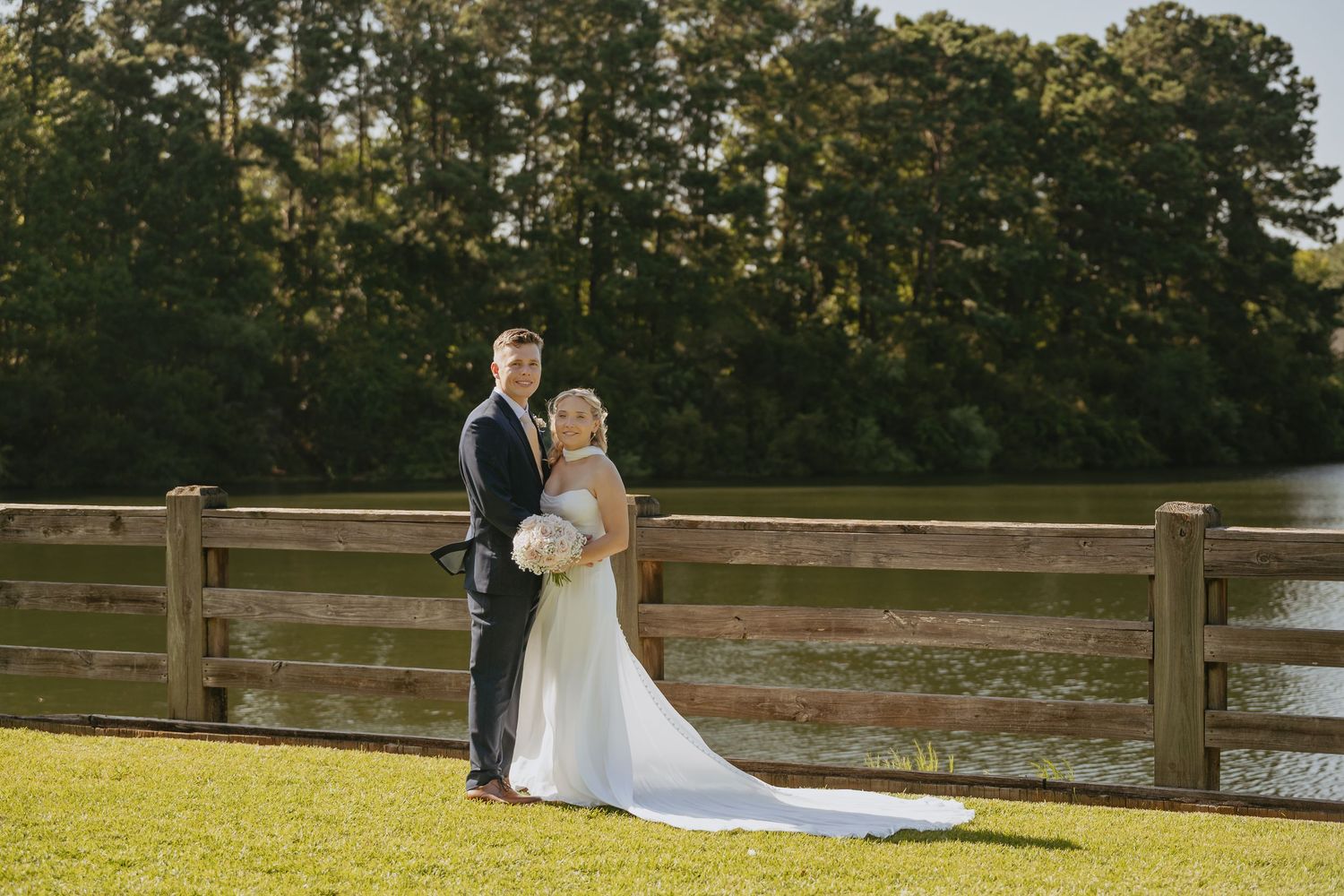 A bride in a long white gown holding a bouquet stands beside her groom in a dark suit at a wooden fence overlooking a peaceful lake.