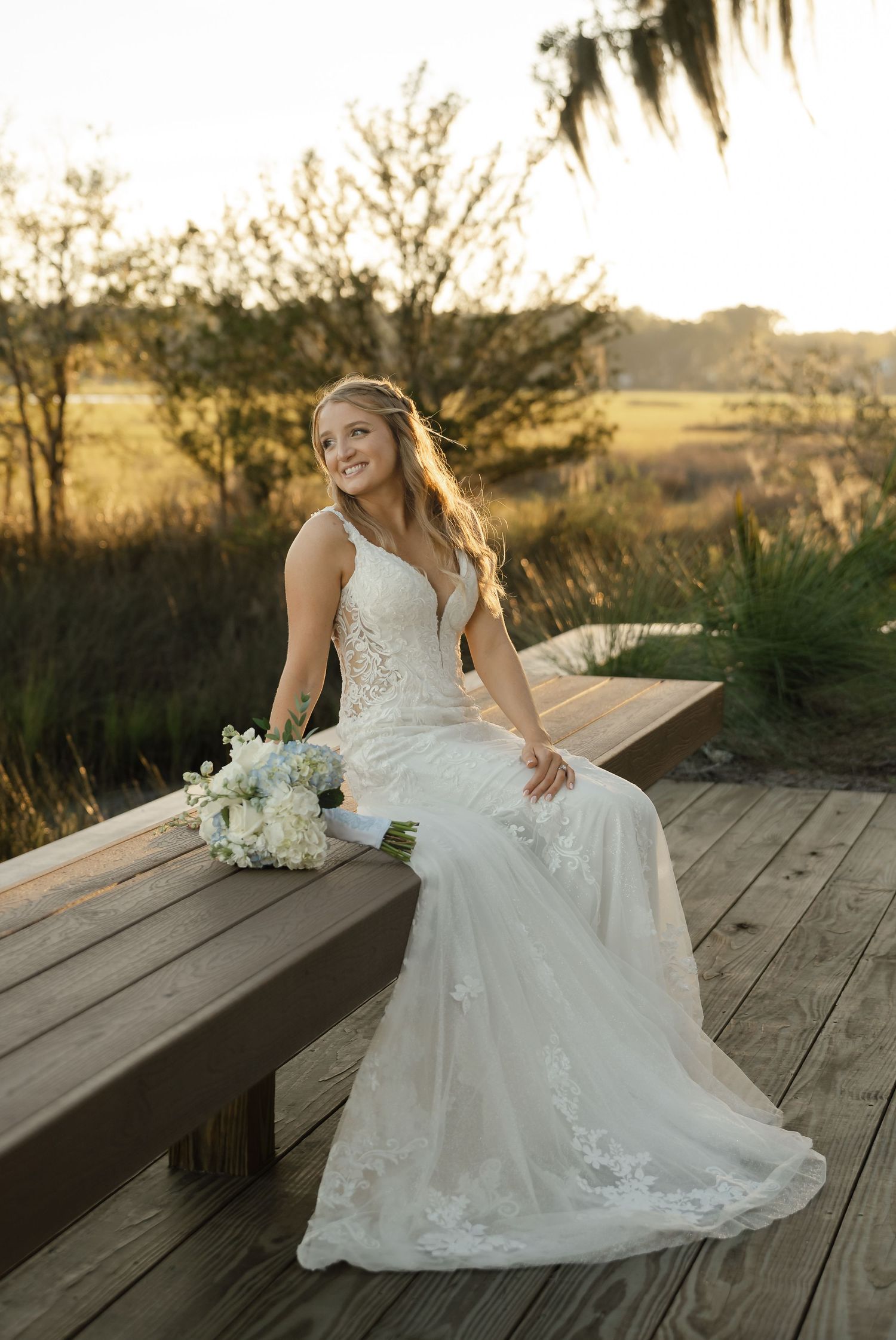 Smiling bride in a lace wedding gown sits on a wooden bench holding a white bouquet in a golden sunset marsh setting.