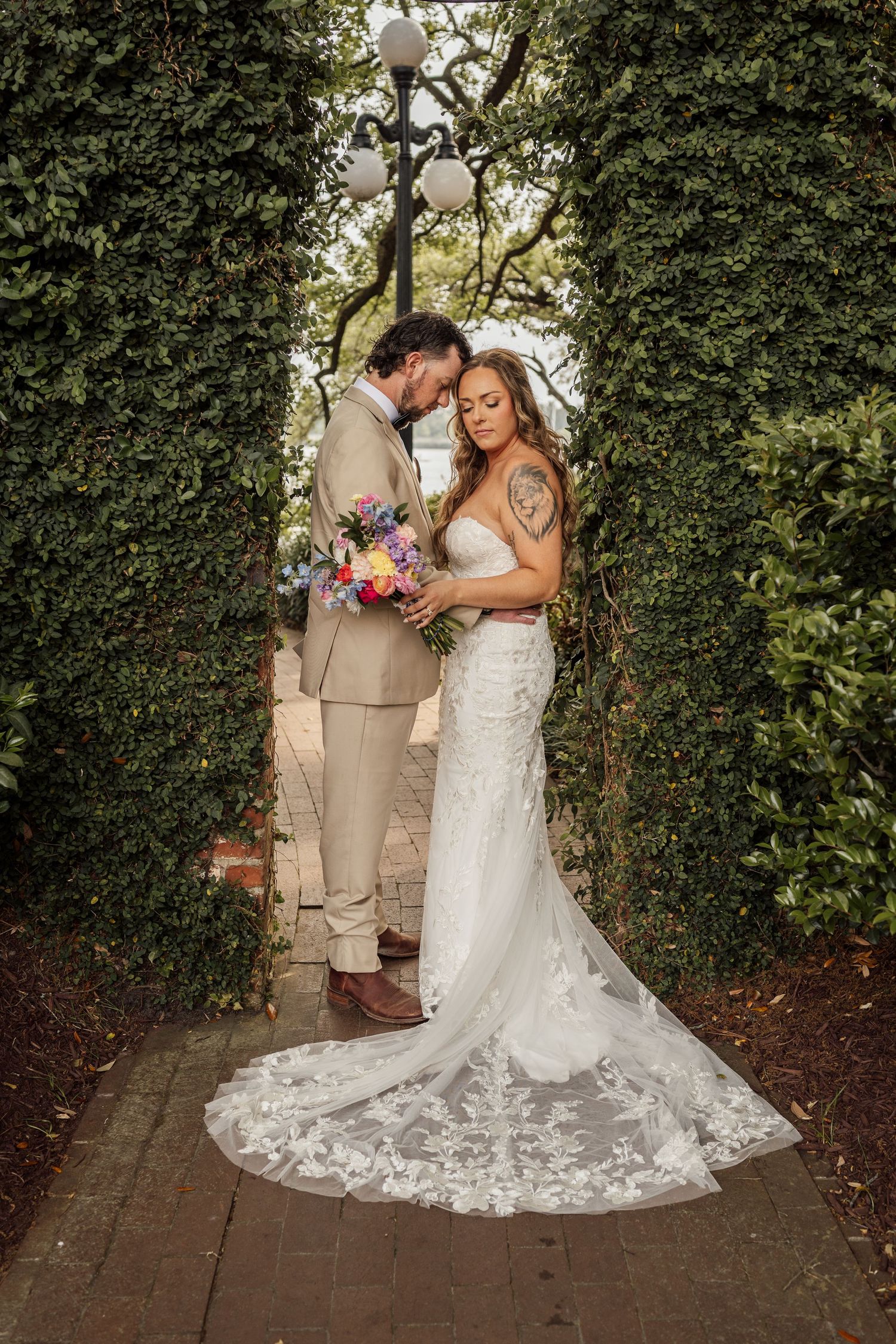 Bride and groom embrace on a brick path between tall ivy hedges, her lace train spread behind and a bright bouquet in hand.