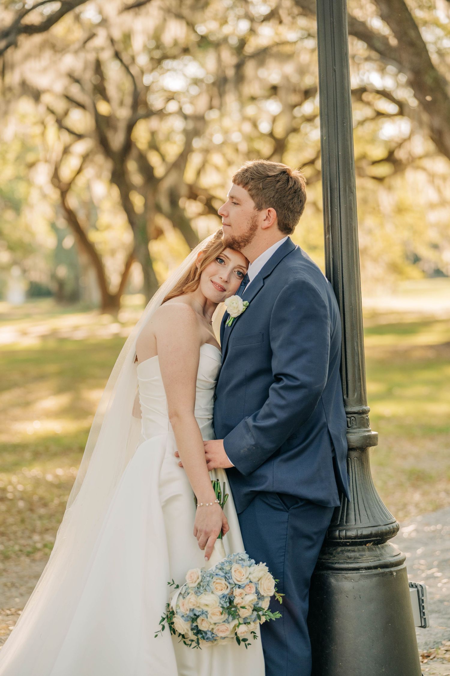 Bride in a strapless white gown and veil rests her head on her groom's chest as they lean against a lamppost holding a bouquet.