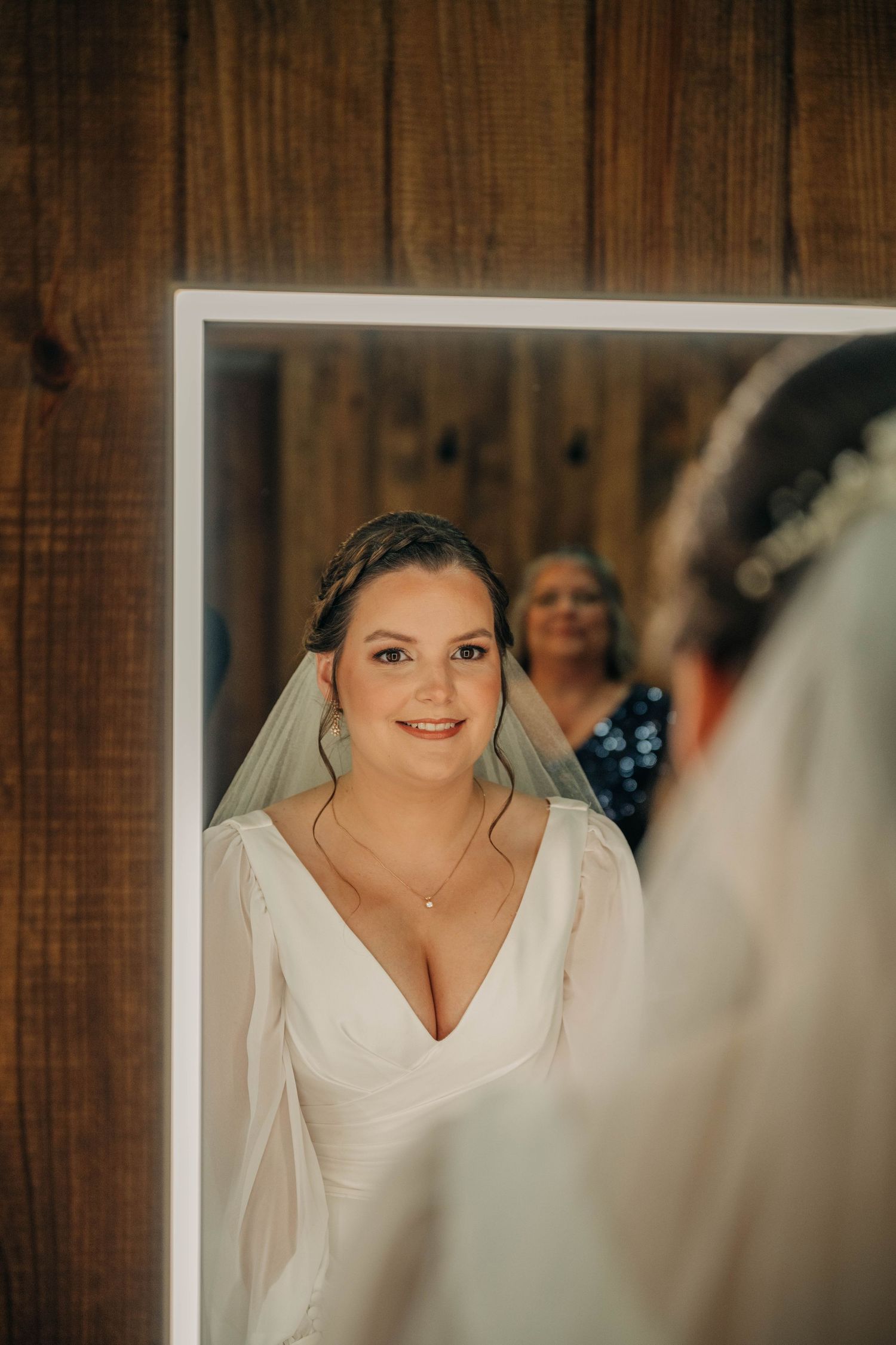 Bride in a veil and V-neck gown smiles at her reflection in a mirror against a rustic wood wall as her mother looks on.
