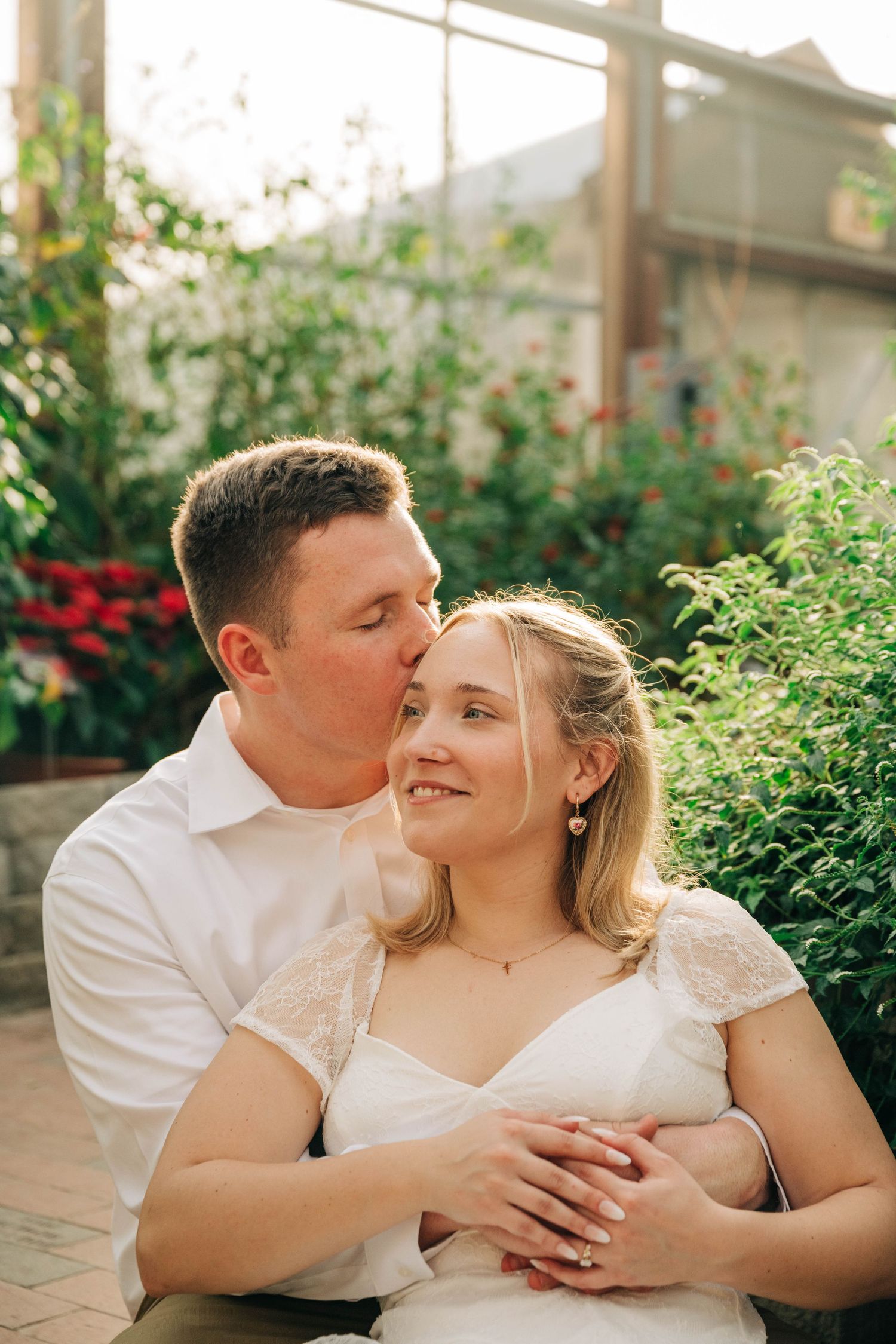 Man in a white shirt kisses his smiling fiancée on the forehead, her engagement ring visible, in a garden with red flowers.