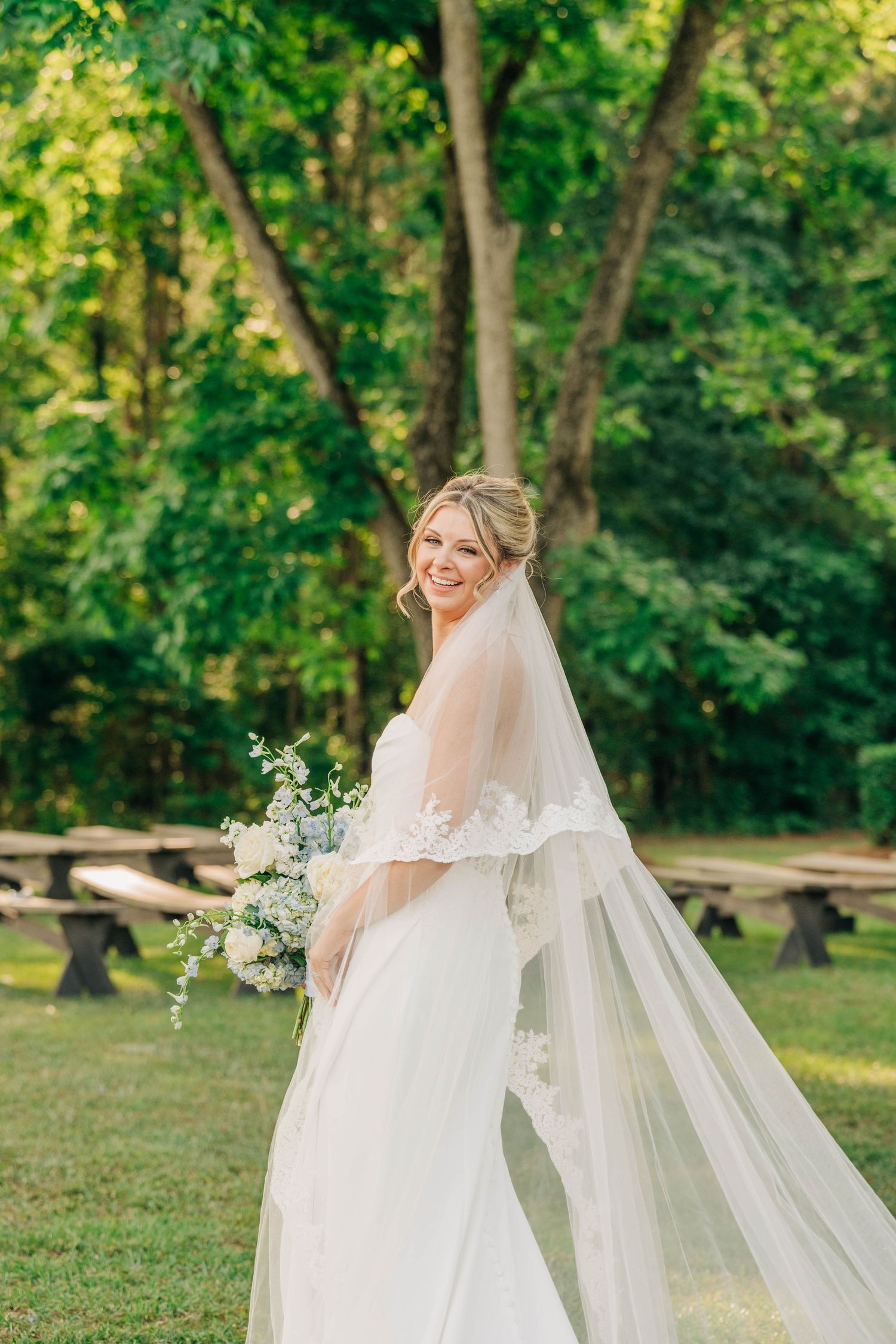 Bride in a white wedding gown with a lace-trimmed cathedral veil smiles over her shoulder holding a flower bouquet outdoors.