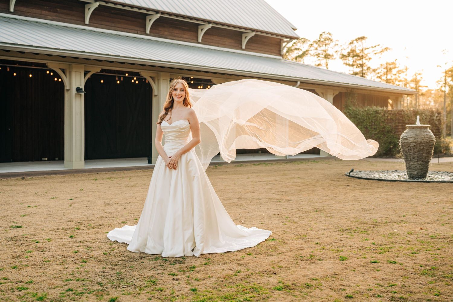 Bride in a strapless wedding gown with a billowing veil poses in golden light outside a rustic venue with a stone fountain.