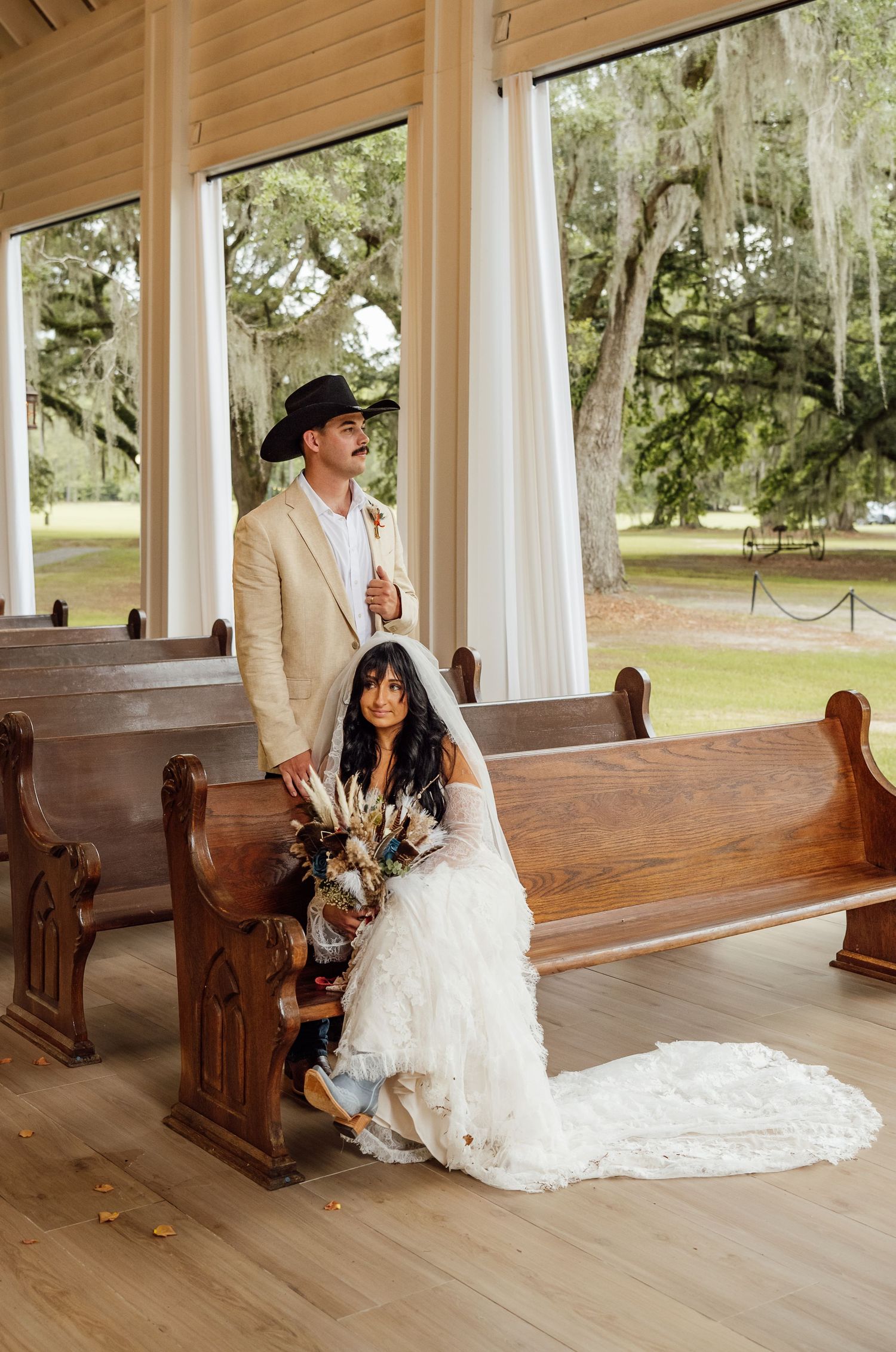 Bride in a ruffled white gown and veil sits on a wooden church pew beside her groom in a tan suit and black cowboy hat.