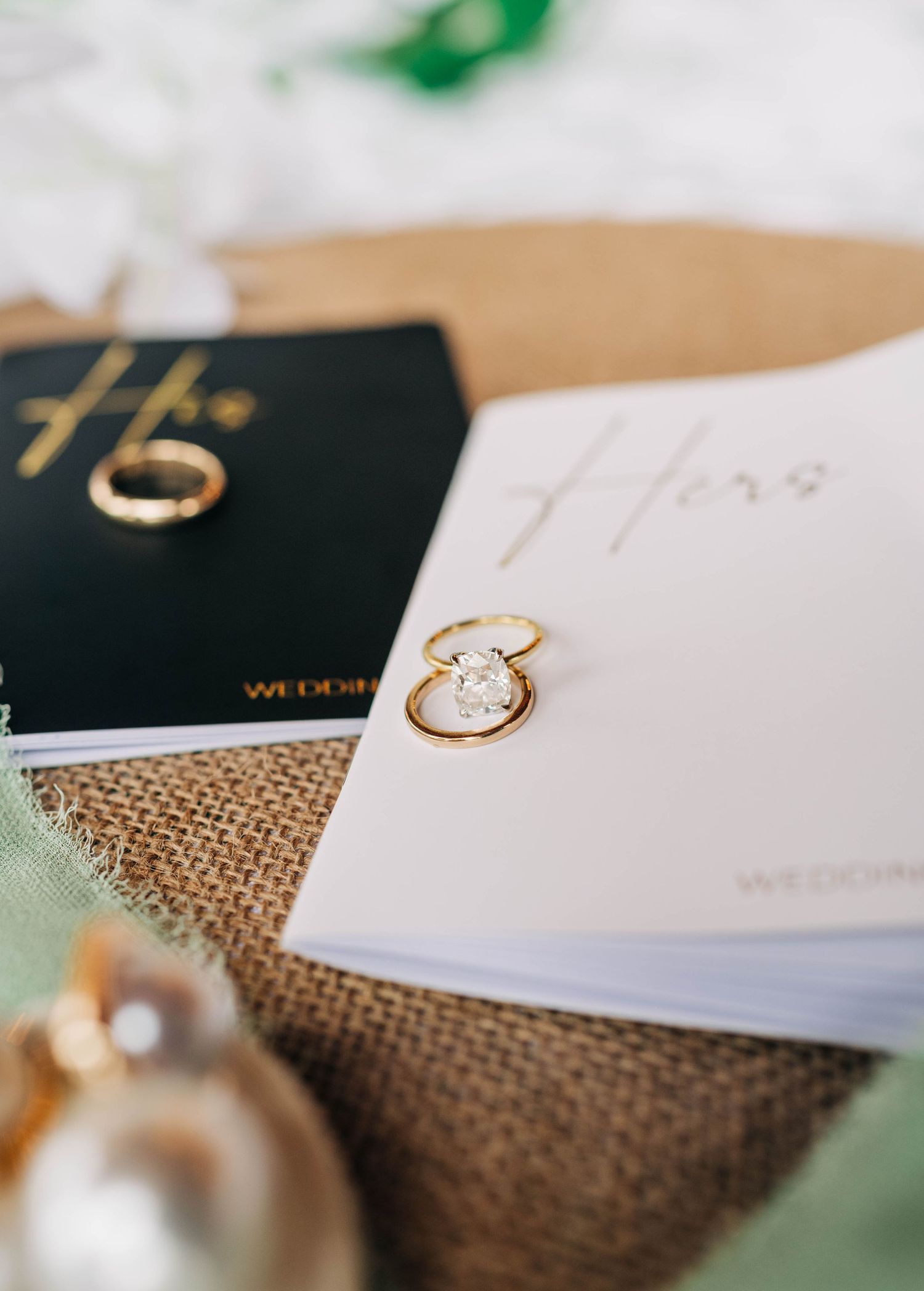 Gold wedding bands and a diamond engagement ring resting on black and white his and hers wedding vow books atop burlap.