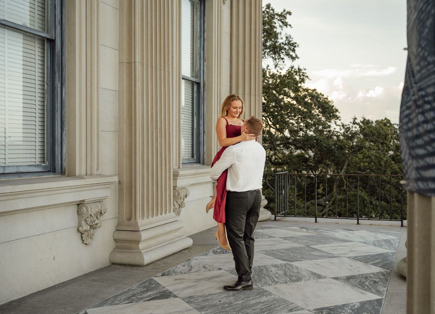 Engaged couple embracing on a patterned stone terrace beside classical columns, the woman wearing a red dress.