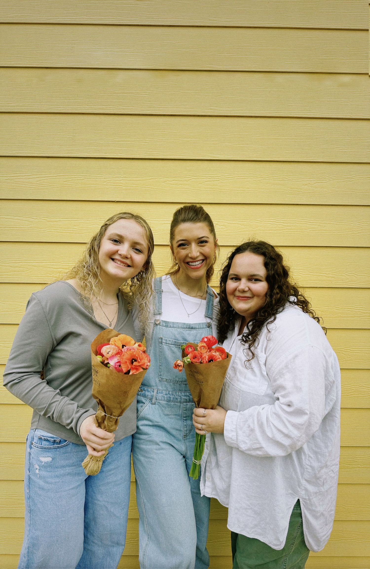 Friends holding bouquets of coral flowers pose against a bright yellow wall.