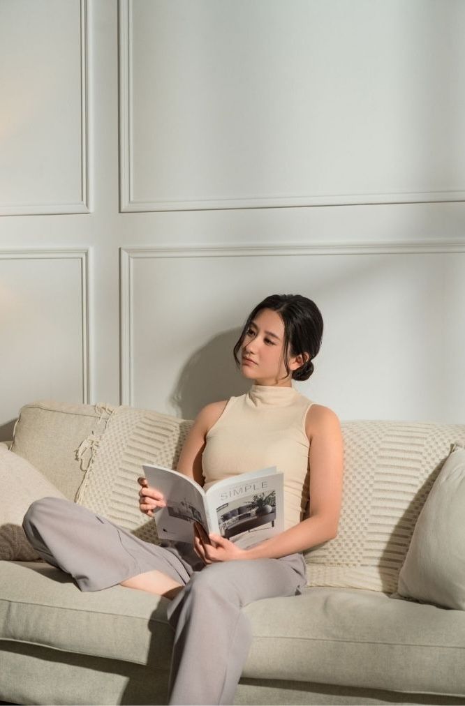 A person in a beige turtleneck and gray pants relaxes on a cream-colored couch while reading a magazine.