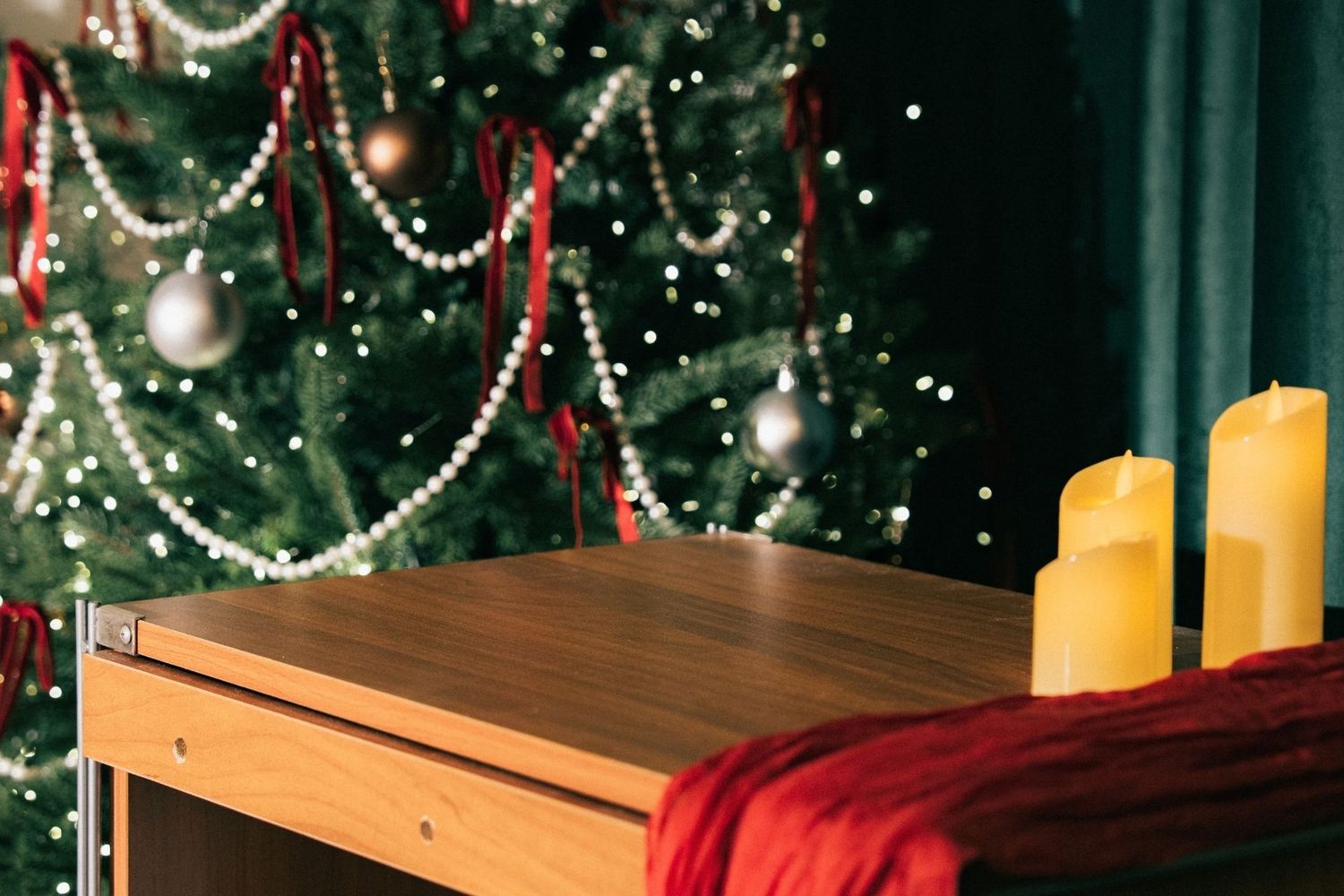 Wooden desk with yellow candles near a decorated Christmas tree with silver baubles and red ribbons in festive setting.