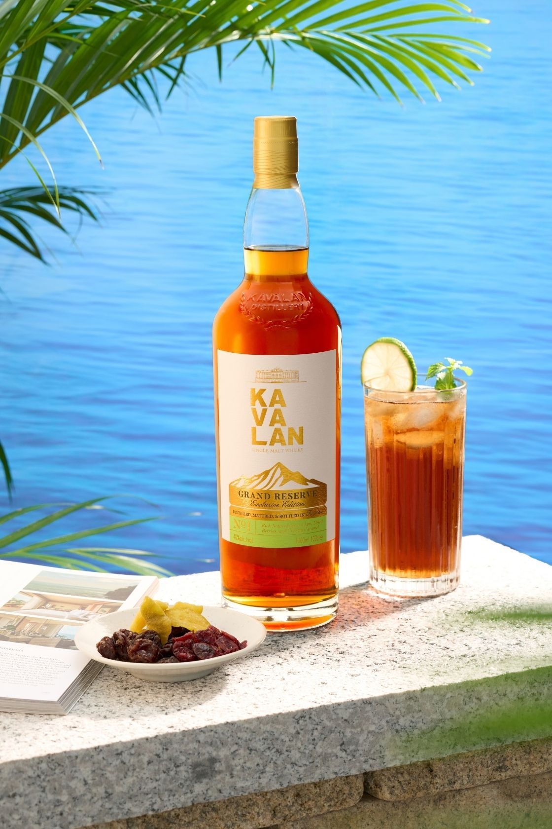 Kavalan Grand Reserve whisky bottle beside a tall iced cocktail with lime and mint, set against a tropical blue ocean backdrop.