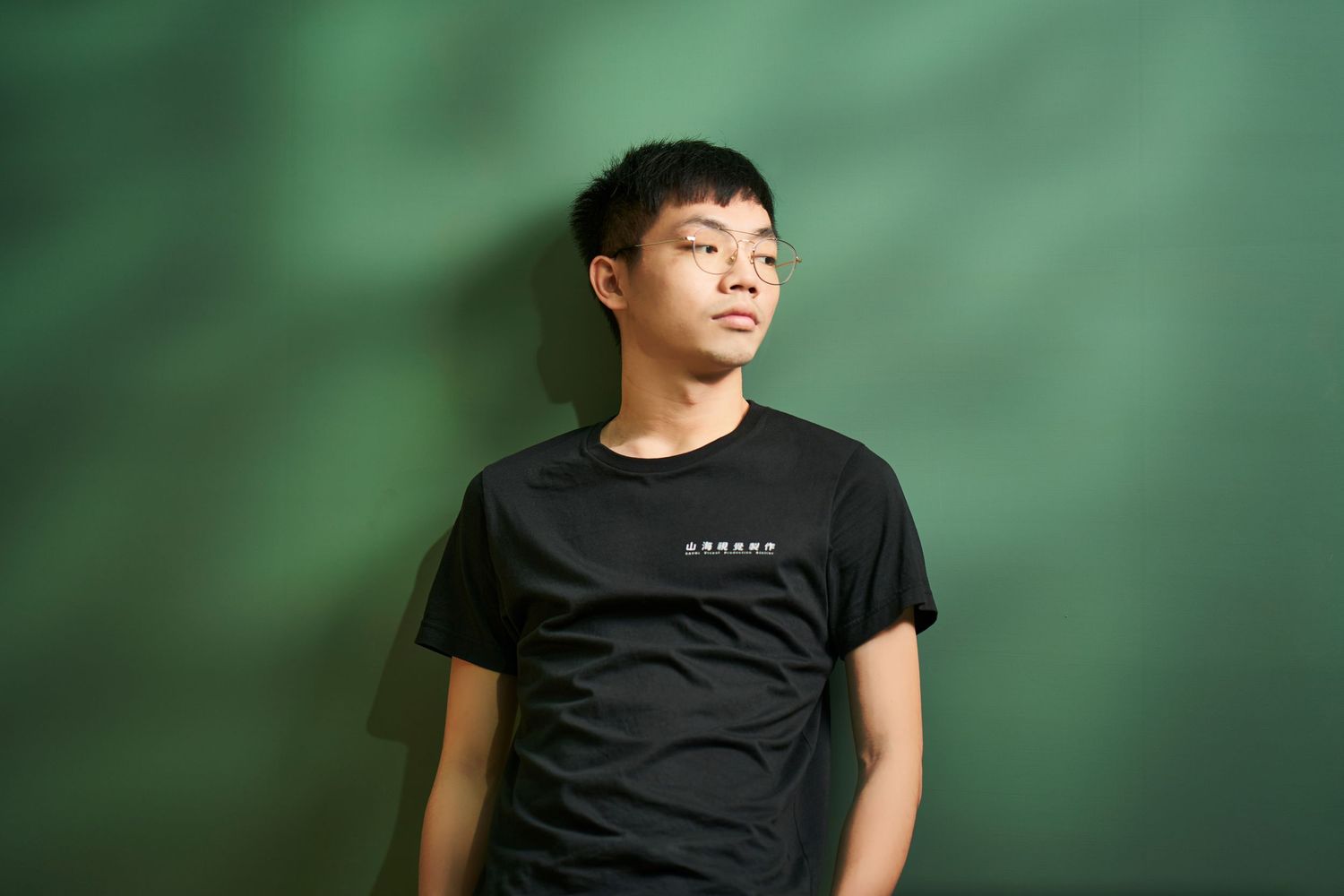 Person wearing a black t-shirt stands against a green background in a casual portrait pose.