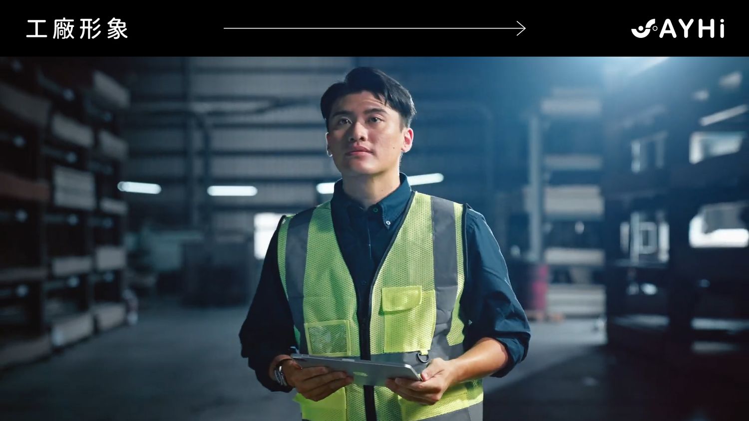 A young male factory worker in a high-visibility vest holds a tablet in a dimly lit industrial warehouse.