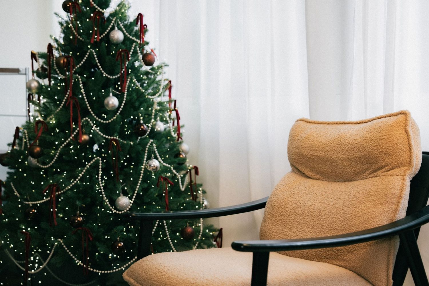Cozy living room with a decorated Christmas tree featuring silver ornaments and garlands next to a plush beige armchair.